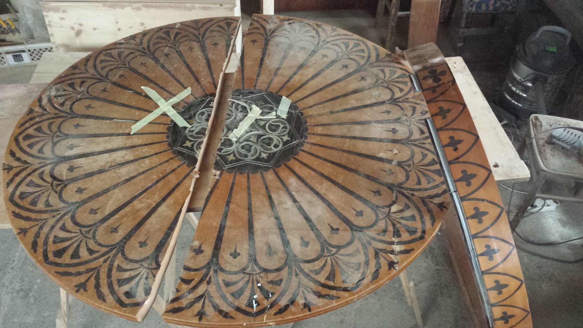Fragmenty okrągłego, drewnianego blatu stołu z dekoracyjnym, radialnym wzorem w kolorach brązu i czerni, zabezpieczone taśmą klejącą podczas renowacji.