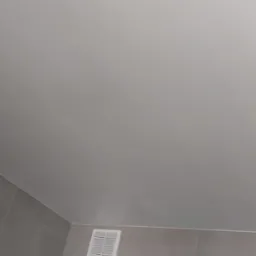 Gładka, jasna powierzchnia sufitu z widoczną kratką wentylacyjną i fragmentem ściany wyłożonej płytkami w odcieniach szarości.