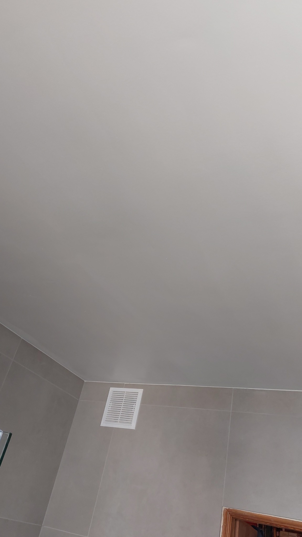 Gładka, jasna powierzchnia sufitu z widoczną kratką wentylacyjną i fragmentem ściany wyłożonej płytkami w odcieniach szarości.