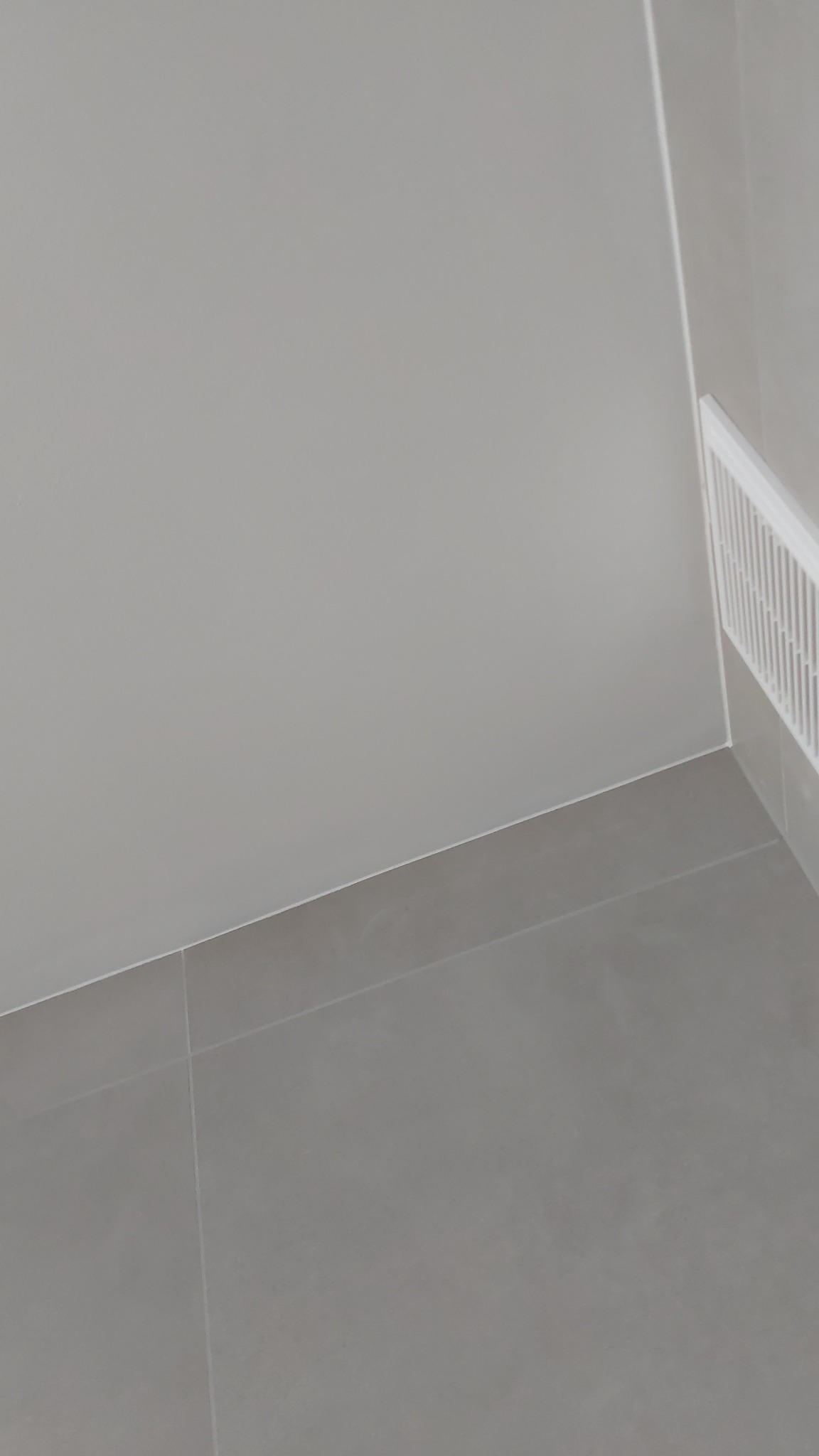 Szare płytki podłogowe z fugami, szara ściana i biała listwa przypodłogowa z kratką wentylacyjną w rogu pomieszczenia.