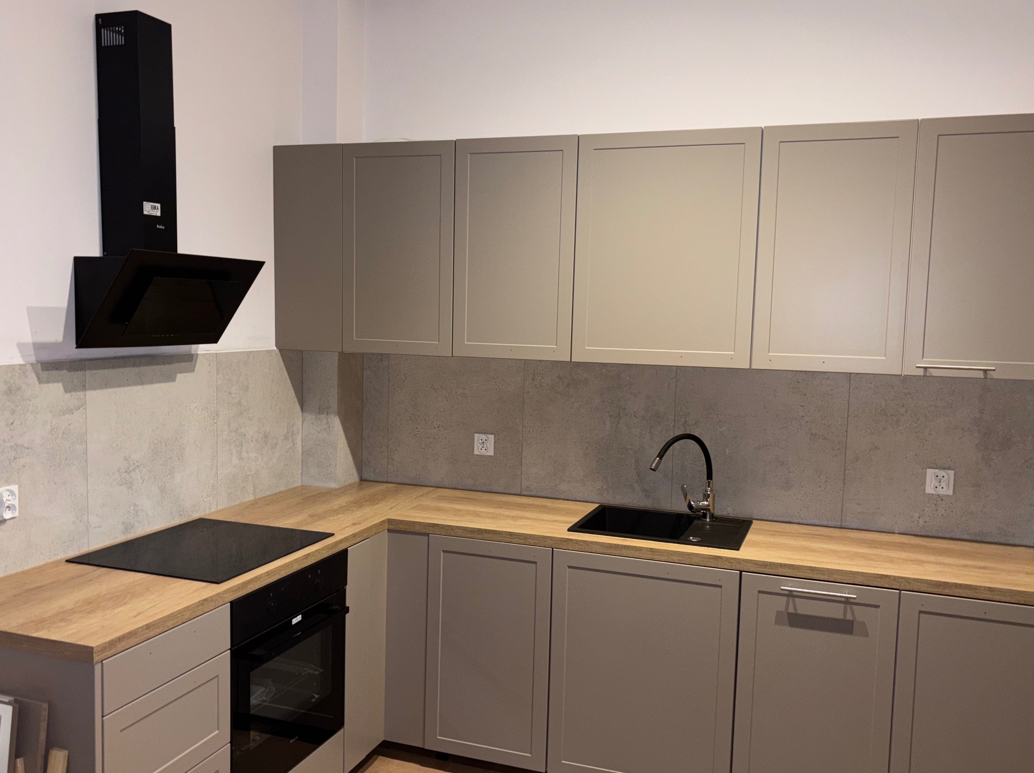 Nowoczesna kuchnia w szarościach z drewnianym blatem. Czarny zlew i płyta indukcyjna. Minimalistyczny okap. Szare płytki na ścianie. Gniazdka elektryczne.