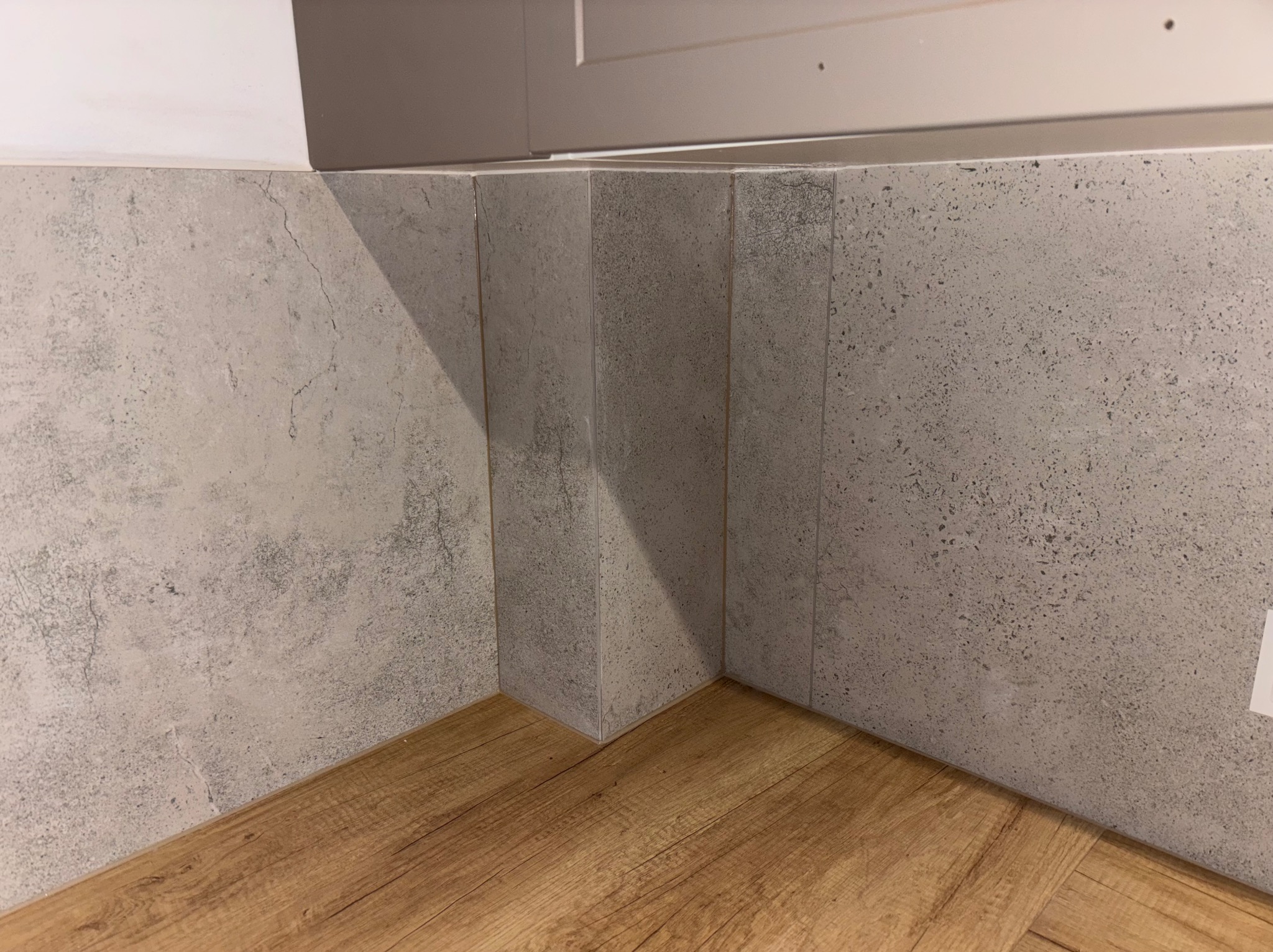 Szare płytki imitujące beton na ścianie przy podłodze z paneli drewnopodobnych, widok z dołu, fragment szafki w tle, fuga w kolorze płytek.