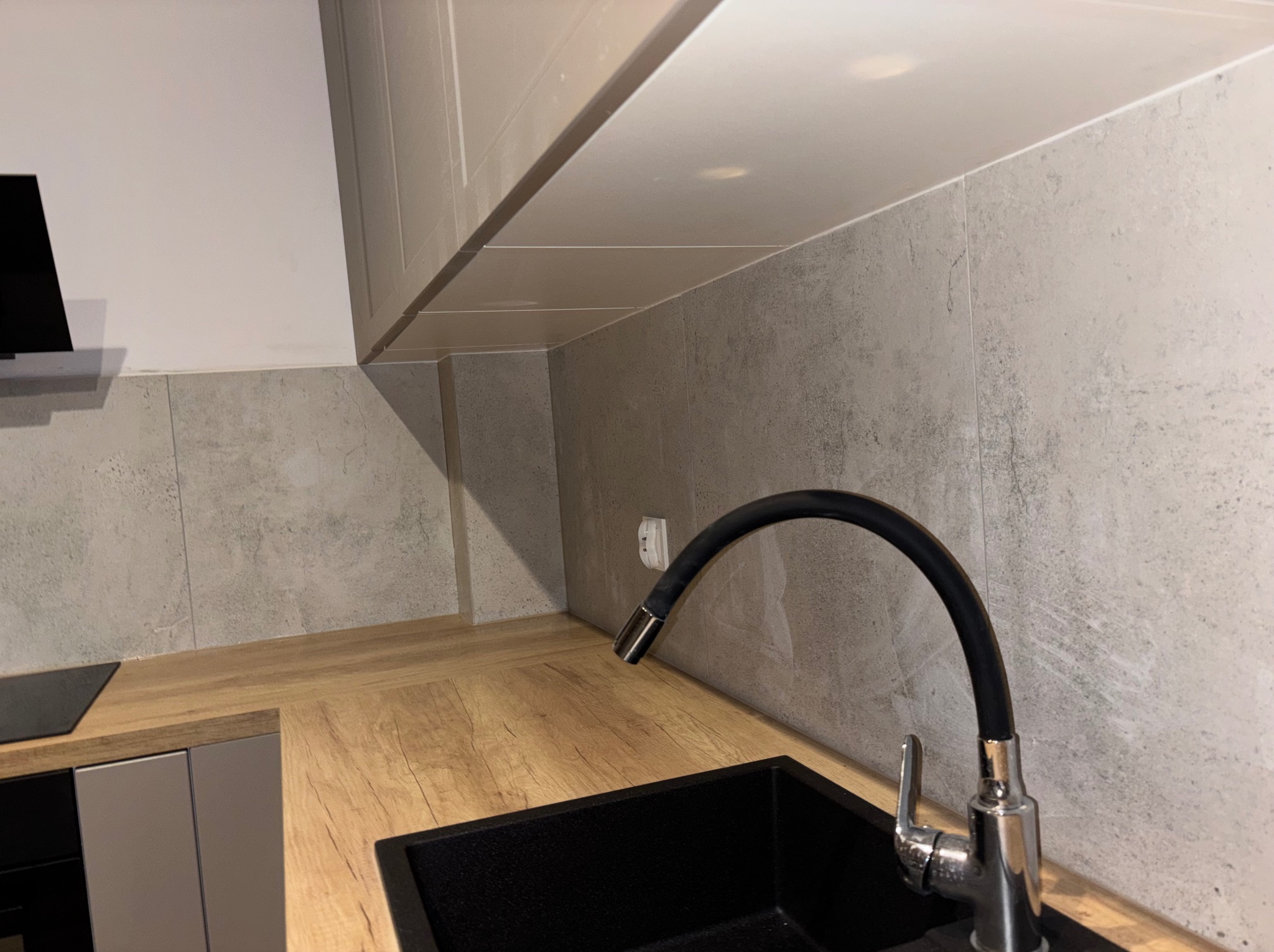 Nowoczesna kuchnia: czarny zlew z chromowaną baterią na tle szarych płytek i drewnianego blatu. Minimalistyczny design, subtelne oświetlenie.