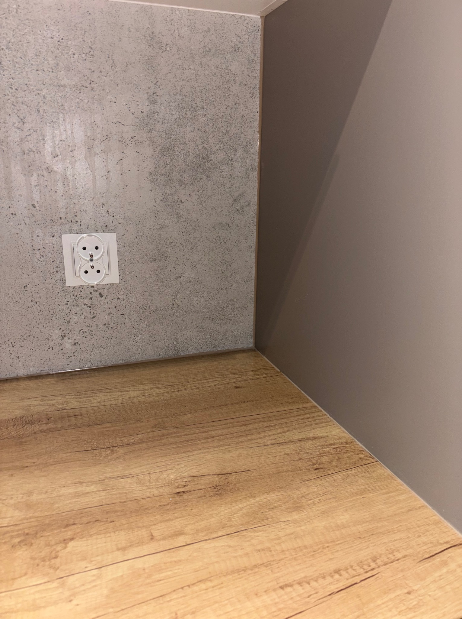 Szara ściana z gniazdkiem, drewniany blat i fragment pomalowanej ściany w odcieniach beżu. Minimalistyczne wykończenie wnętrza, detale aranżacji.