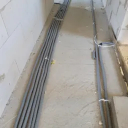 Szereg szarych, karbowanych rur ochronnych na przewody elektryczne ułożonych wzdłuż surowej ściany z betonu kom&oacute;rkowego w trakcie prac instalacyjnych, zabezpieczonych białymi opaskami zaciskowymi.