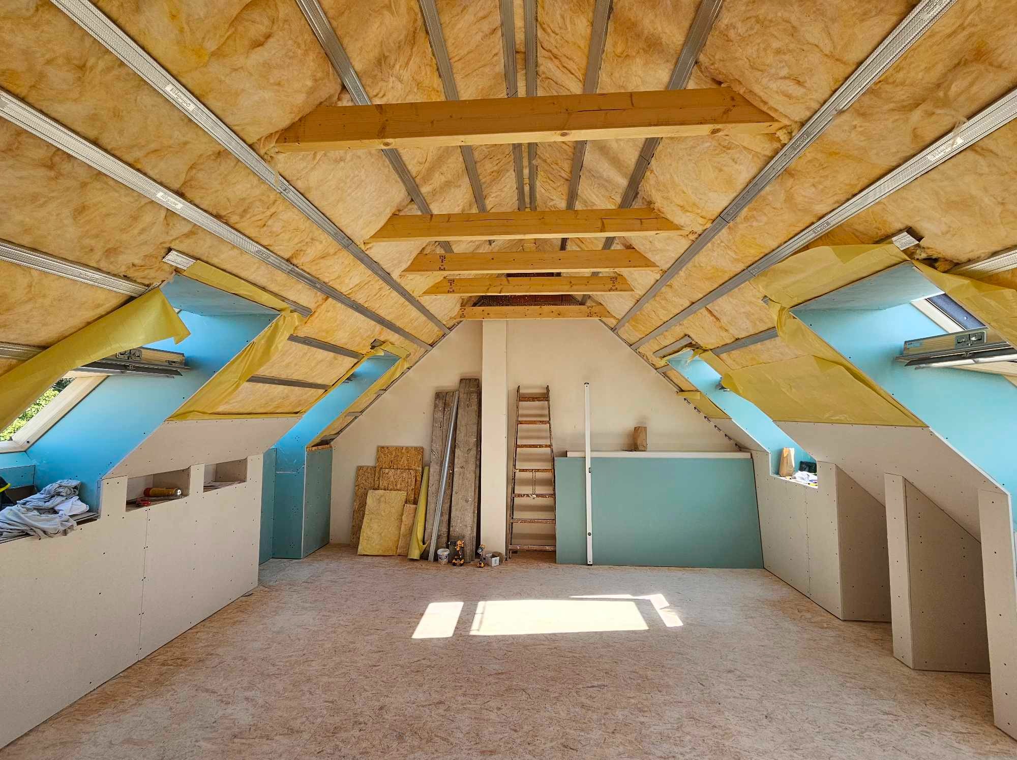 Poddasze w trakcie adaptacji: widoczne ocieplenie wełną mineralną, metalowe profile, drewniane belki, częściowo zamontowane płyty na ścianach i skosach, oraz drewniana podłoga OSB.