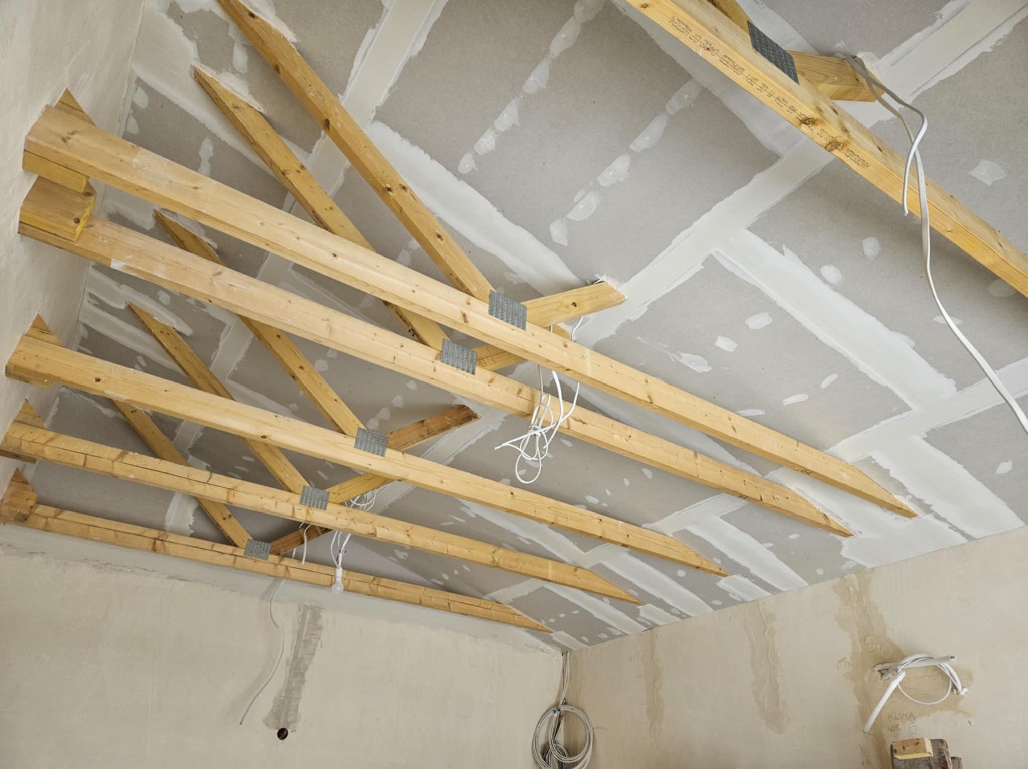 Sufit poddasza z widoczną drewnianą konstrukcją więźby dachowej i fragmentami płyt gipsowo-kartonowych, spoiny wypełnione masą szpachlową, zwisające kable elektryczne, surowy stan wykończenia.