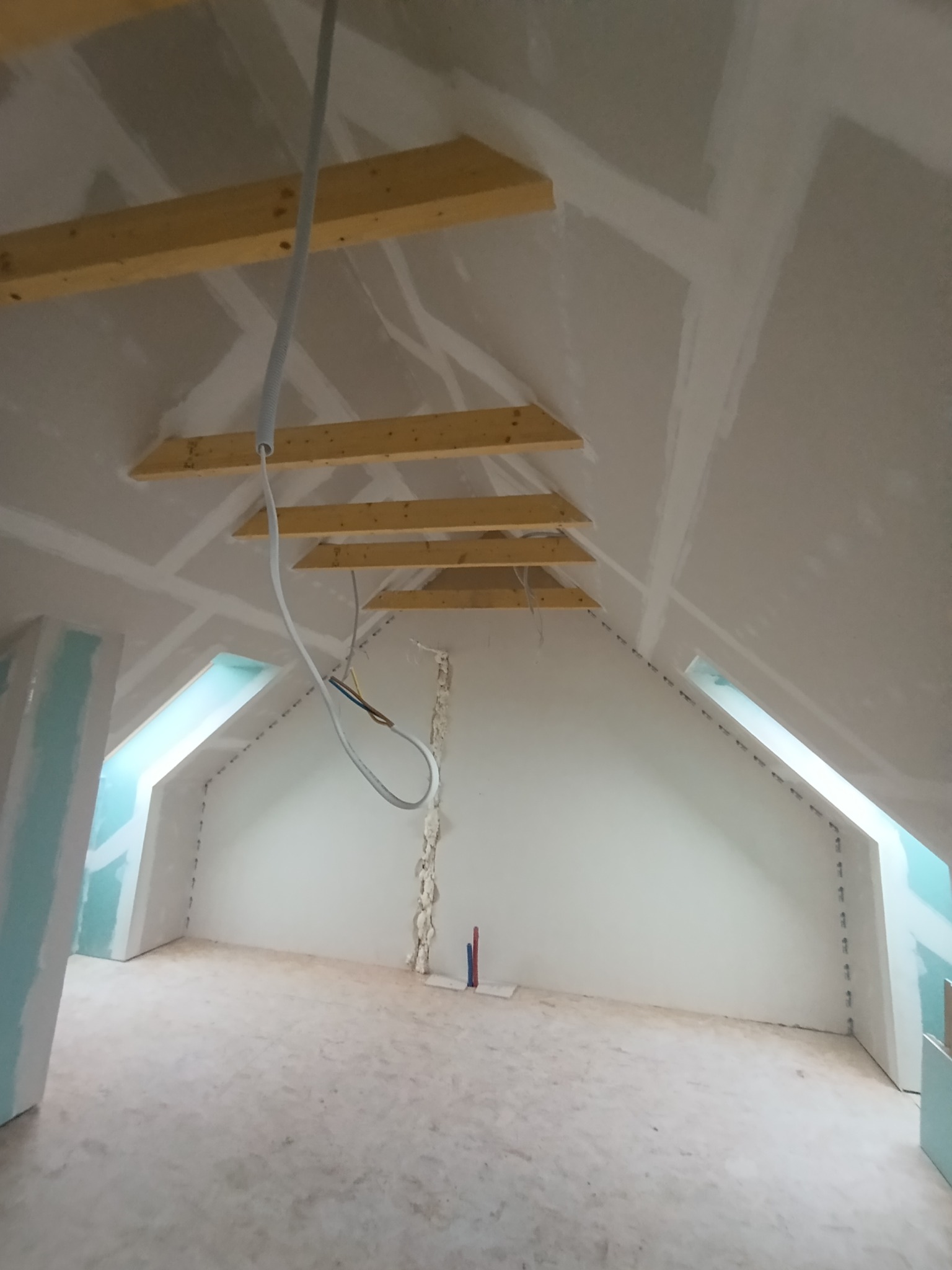 Poddasze w trakcie prac wykończeniowych: widoczne drewniane elementy konstrukcyjne, częściowo zamontowane płyty, przewody elektryczne zwisające z sufitu, oraz ślady pianki montażowej wokół rur.