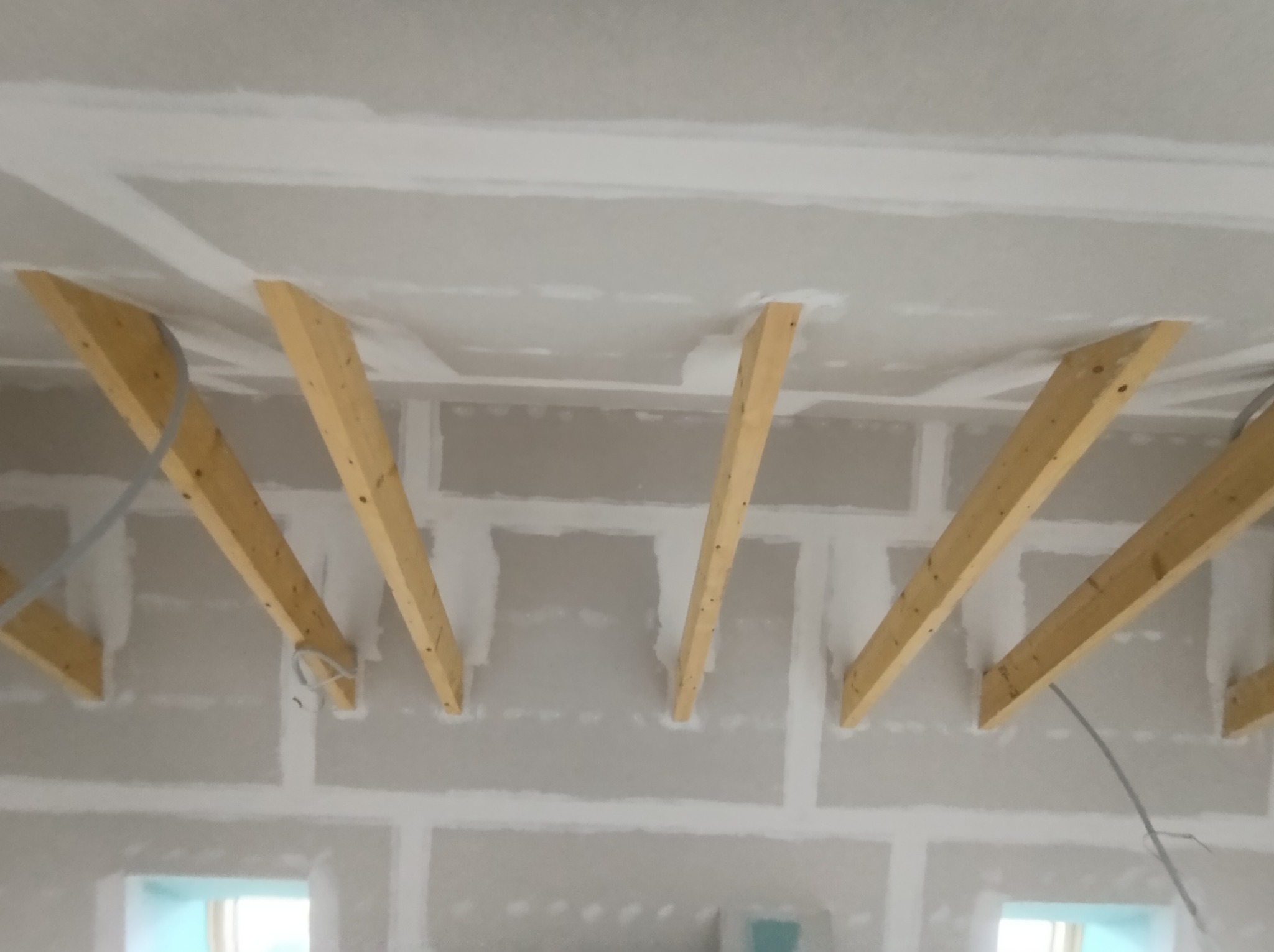 Sufit i ściany w trakcie montażu, widoczne drewniane belki konstrukcyjne i płyty gipsowo-kartonowe z widocznymi łączeniami przed wykończeniem, przewody elektryczne zwisają z belek.