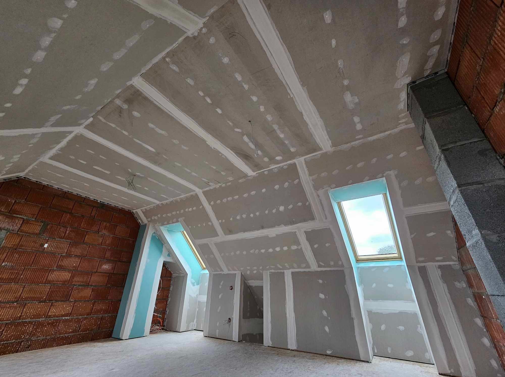 Poddasze z oknami dachowymi w trakcie wykańczania płytami karton-gips, widoczne łączenia płyt zaszpachlowane, ściana z czerwonej cegły w lewym rogu.
