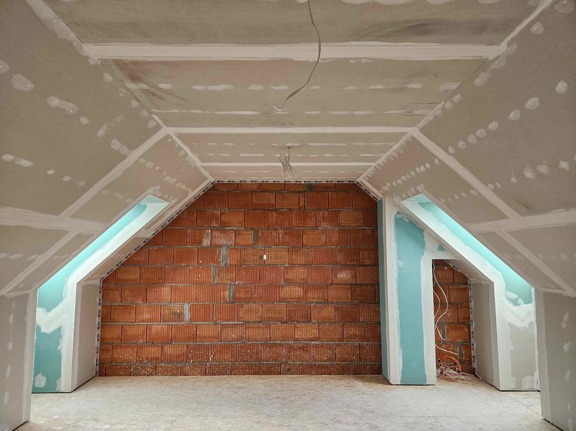 Poddasze w trakcie adaptacji: widoczne cegły, konstrukcja z płyt z widocznymi łączeniami, surowe kable zwisające z sufitu, przygotowane otwory okienne.