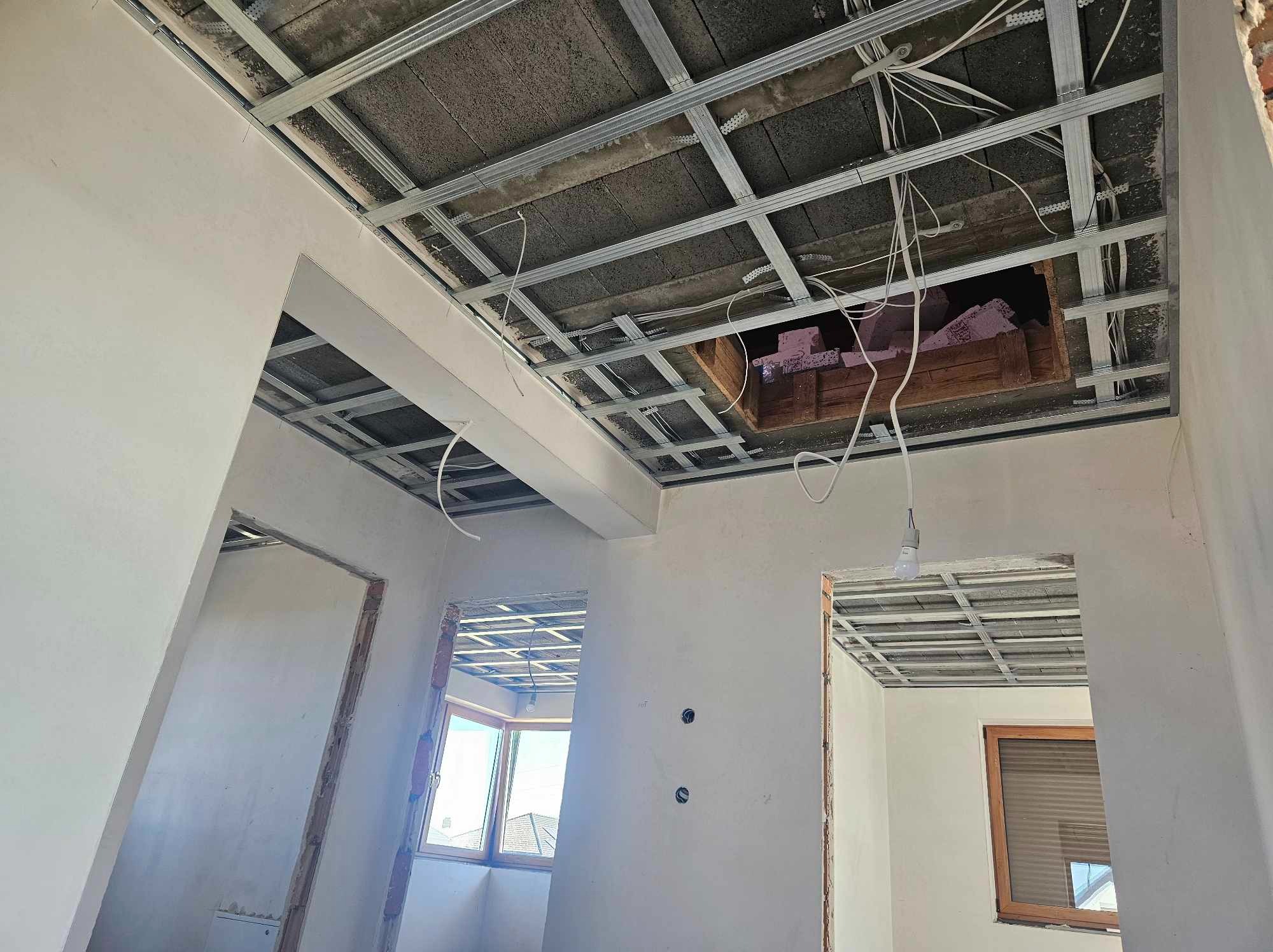 Wnętrze w trakcie prac wykończeniowych: widoczne metalowe profile sufitowe, instalacja elektryczna, fragment ocieplenia w otworze stropowym oraz wstępnie przygotowane ściany z widocznymi puszkami...
