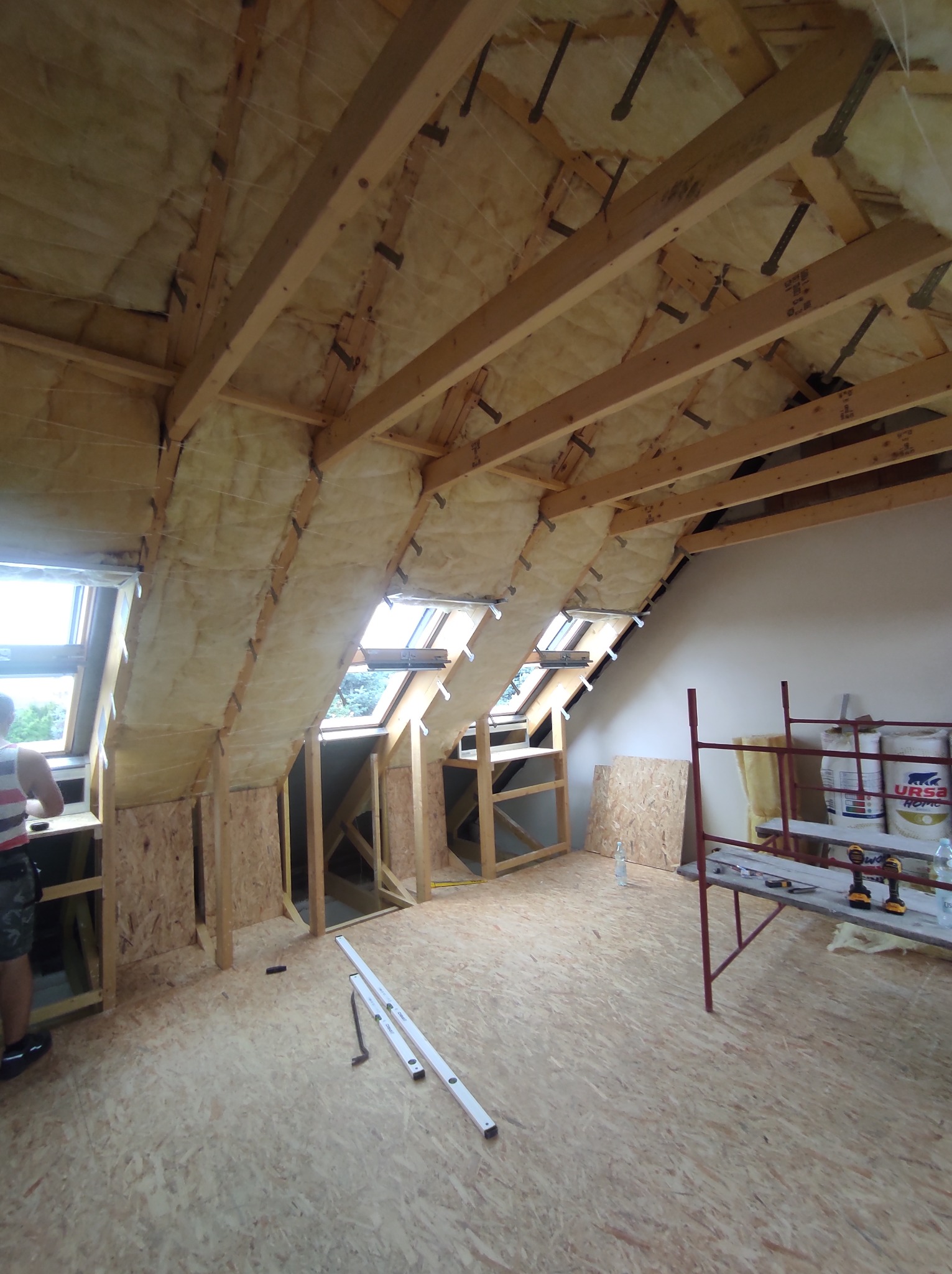 Poddasze w trakcie prac izolacyjnych: widoczne drewniane belki konstrukcyjne, wełna mineralna między nimi oraz okna dachowe. Na podłodze płyty OSB, rusztowanie z narzędziami i worki z materiałami...