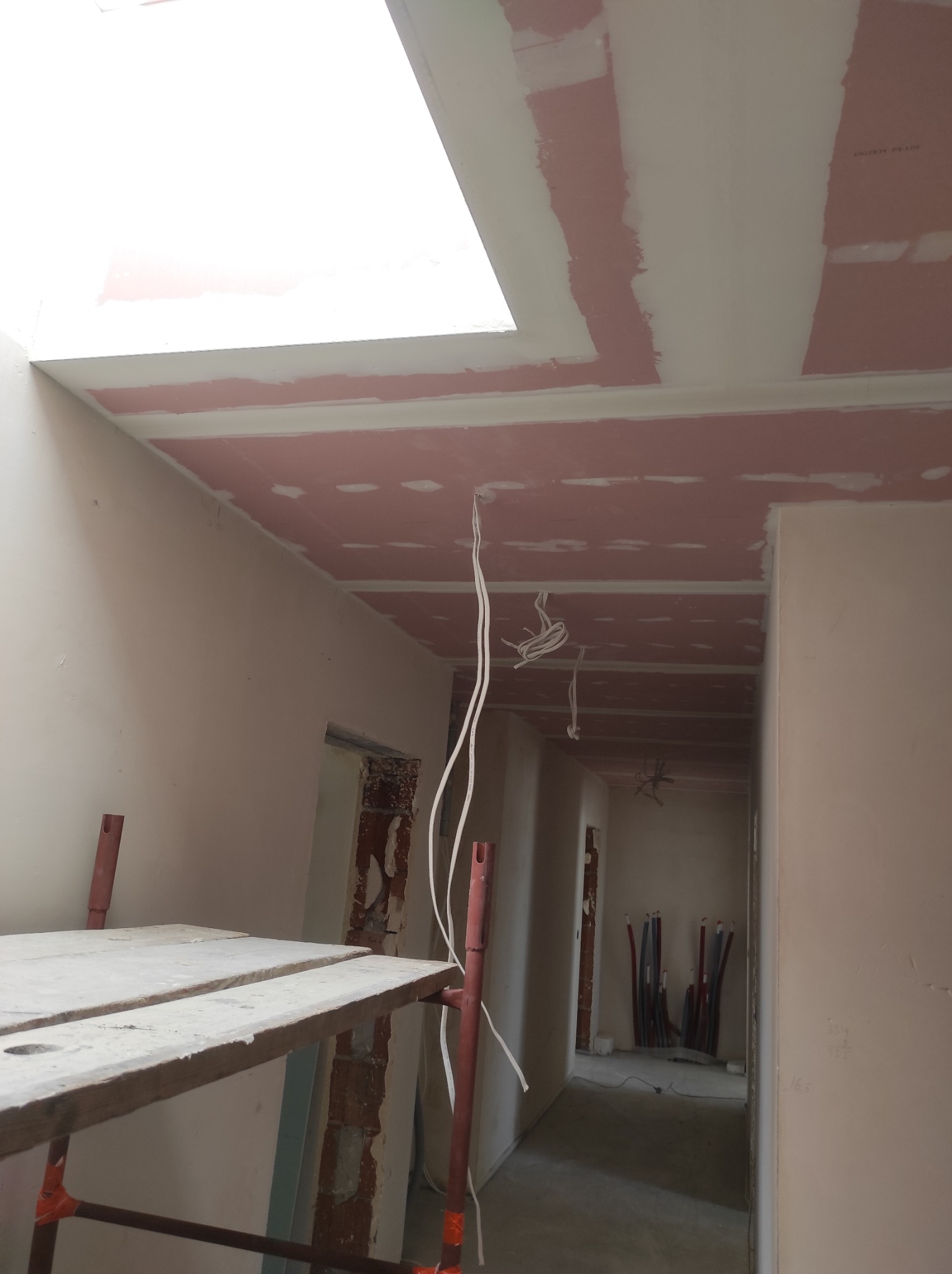 Montaż płyt na suficie podwieszanym z widocznym oknem dachowym, częściowo zaszpachlowane łączenia, widoczne przewody elektryczne, rusztowanie w rogu pomieszczenia, w tle instalacja elektryczna...