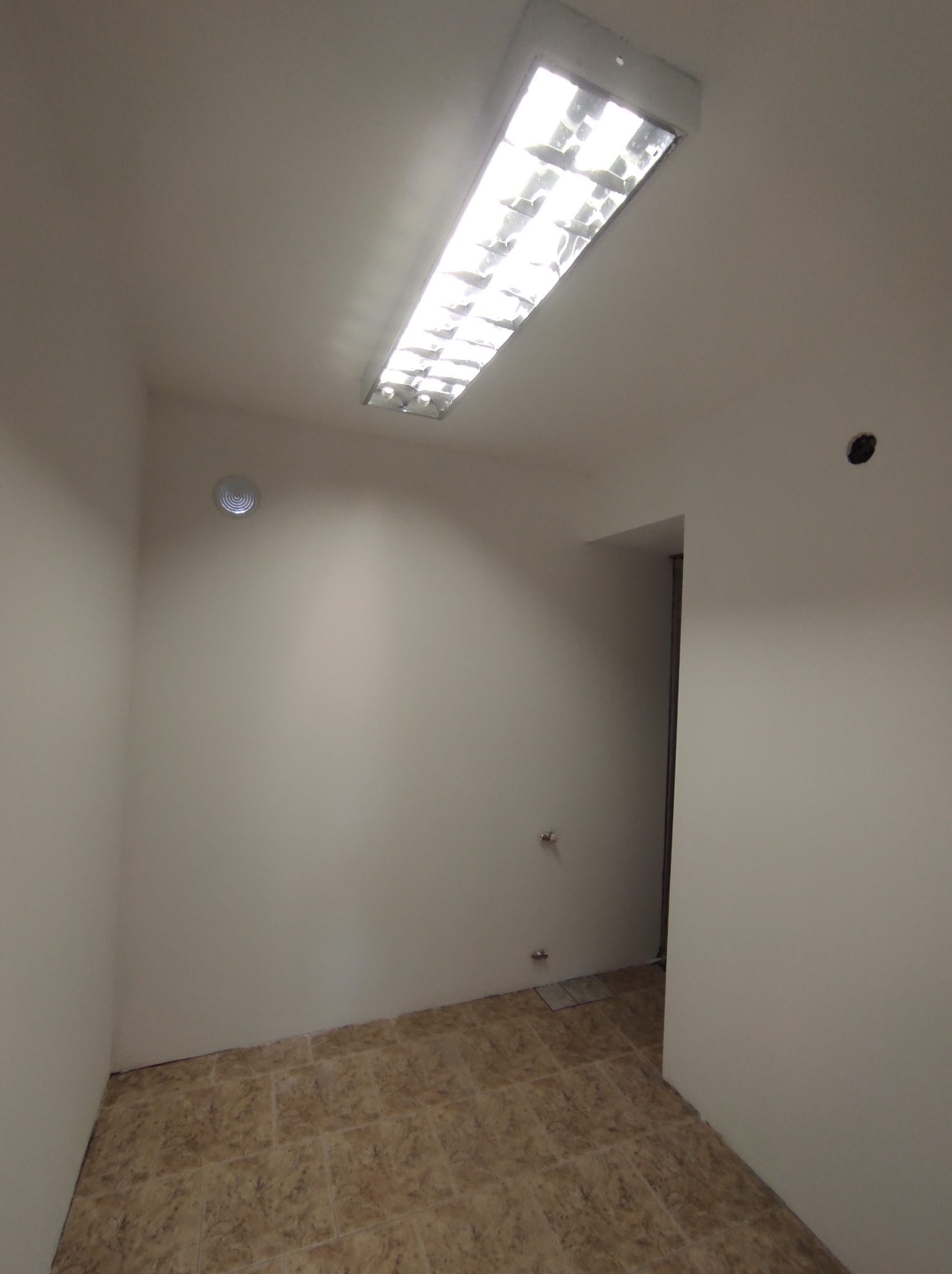 Pomalowane na biało puste pomieszczenie z brązową podłogą z płytek, widoczne otwory w ścianie i jasna lampa sufitowa.