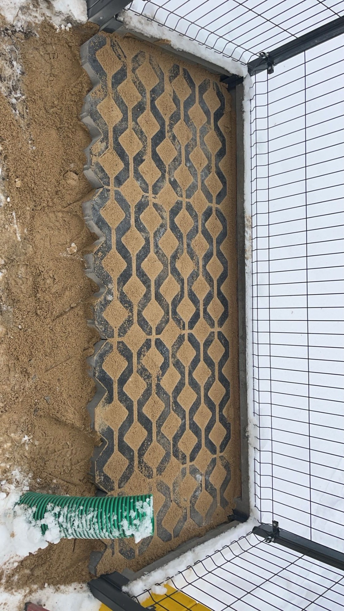 Częściowo wypełnione piaskiem ażurowe płyty betonowe z geometrycznym wzorem, ułożone obok ogrodzenia z siatki, widoczny fragment zielonej rury drenażowej i resztki śniegu.