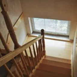 Widok z góry na jasne, drewniane schody z balustradą, prowadzące do okna z widokiem na zieleń.