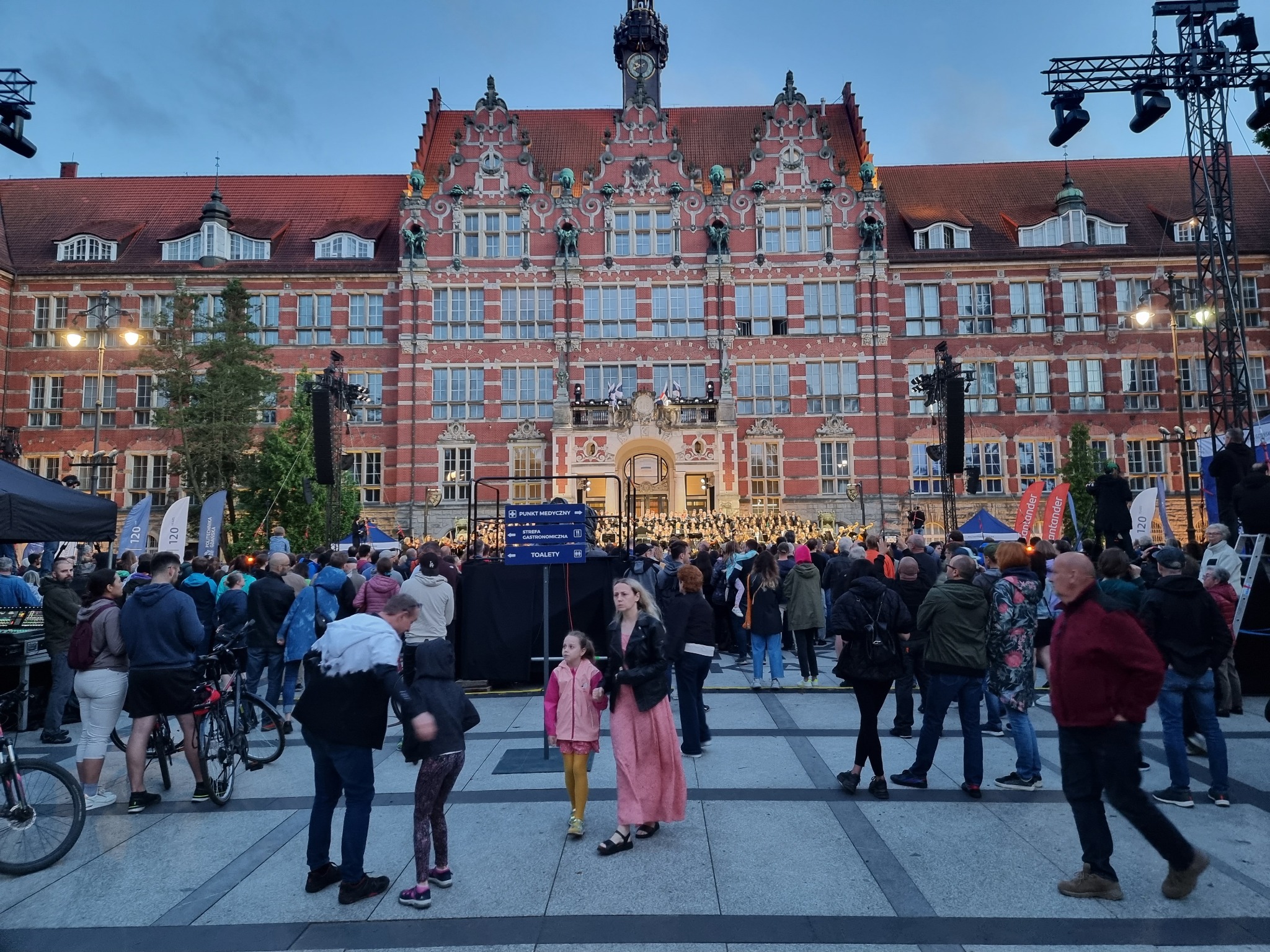 Koncert plenerowy przed budynkiem Politechniki Gdańskiej: tłum ludzi, scena z nagłośnieniem i oświetleniem, tablice informacyjne z napisem 'Toalety'.
