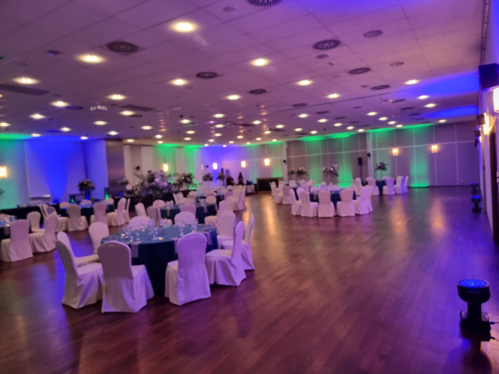 Uroczysta sala balowa z parkietem, oświetlona niebieskim i zielonym światłem, stoły z białymi krzesłami i dekoracjami kwiatowymi, widoczny sprzęt oświetleniowy na podłodze.