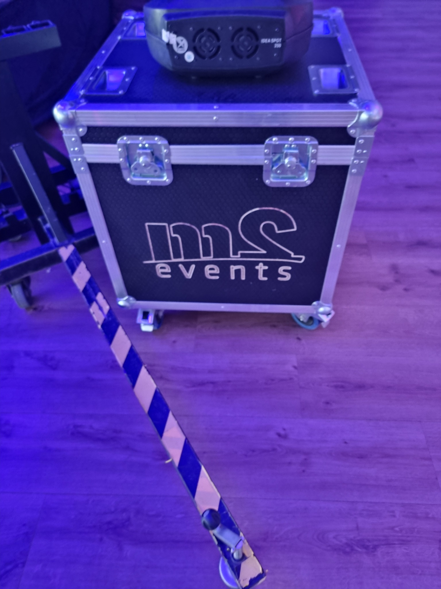 Skrzynia transportowa z logo 'ms events' na kółkach, na niej leży reflektor IDEA SPOT 300, obok stoi statyw oświetleniowy, wszystko w fioletowym oświetleniu.