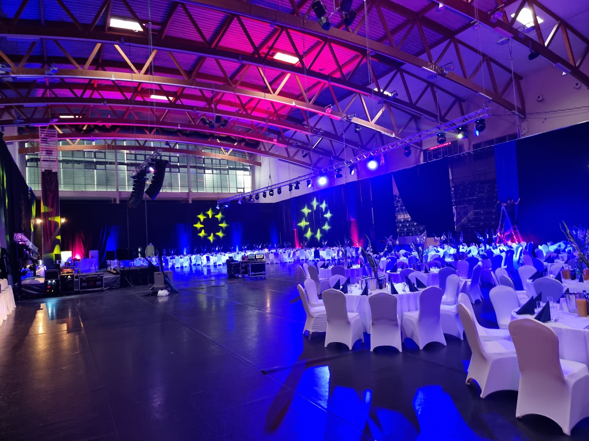 Sala bankietowa przygotowana na galę, oświetlona niebieskim i różowym światłem, z dekoracjami w postaci gwiazd i rzędami stołów z białymi krzesłami.