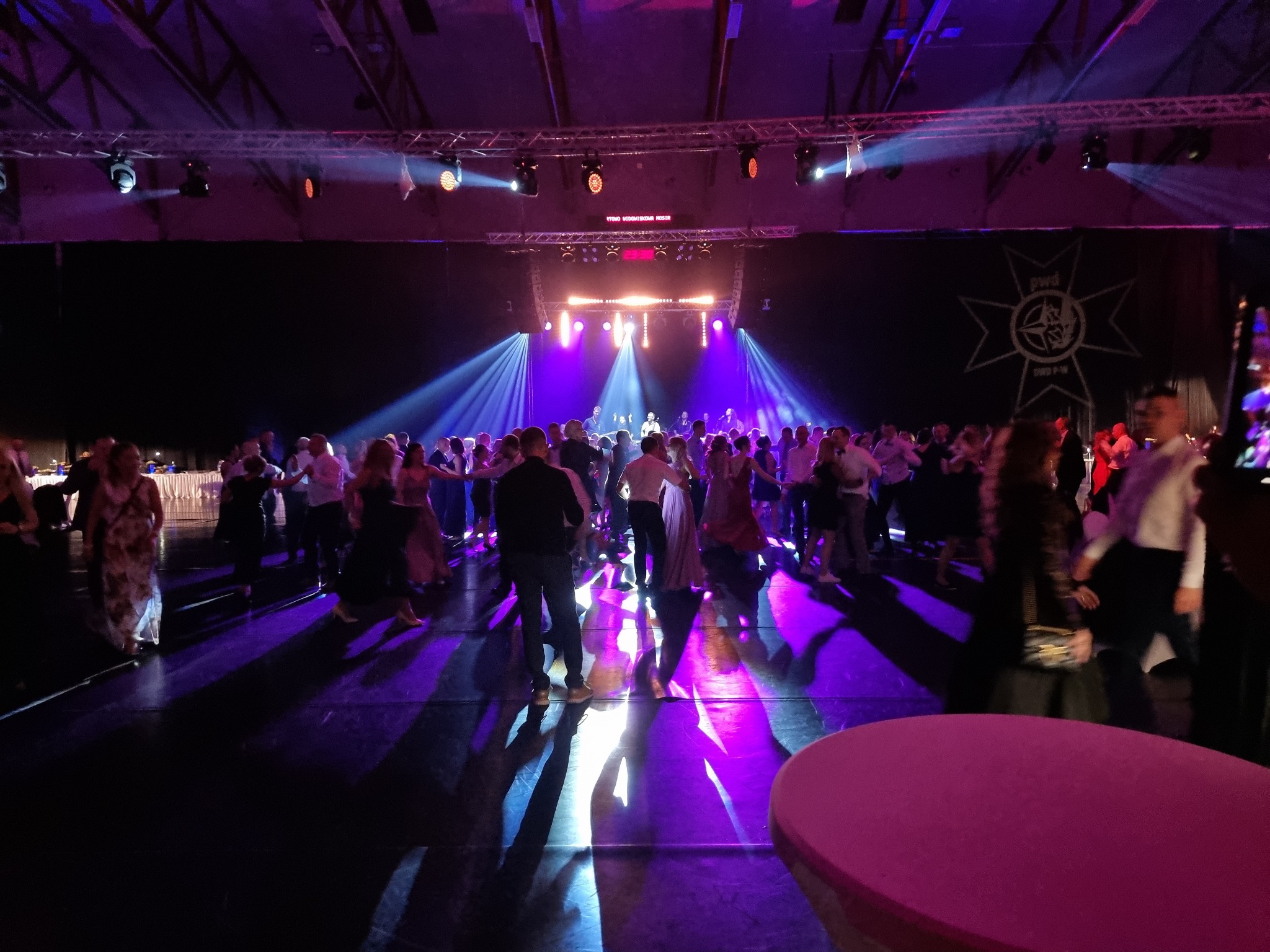 Uczestnicy balu tańczący na parkiecie oświetlonym przez reflektory sceniczne z fioletową poświatą, widoczna scena w tle i logo na ścianie.