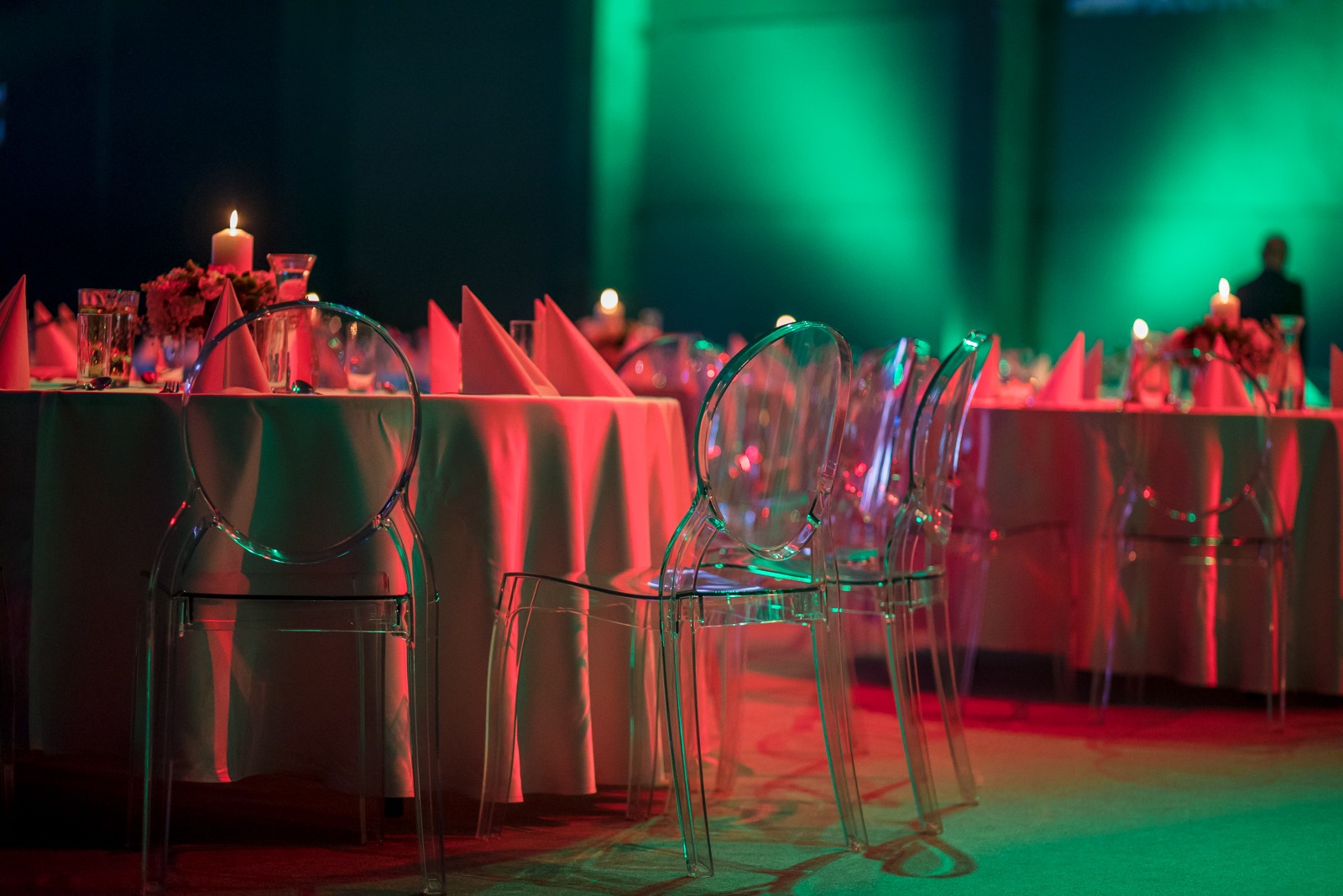 Uroczysta sala bankietowa z okrągłymi stołami, nakryciami, świecami i przezroczystymi krzesłami, oświetlona zielonym i czerwonym światłem.
