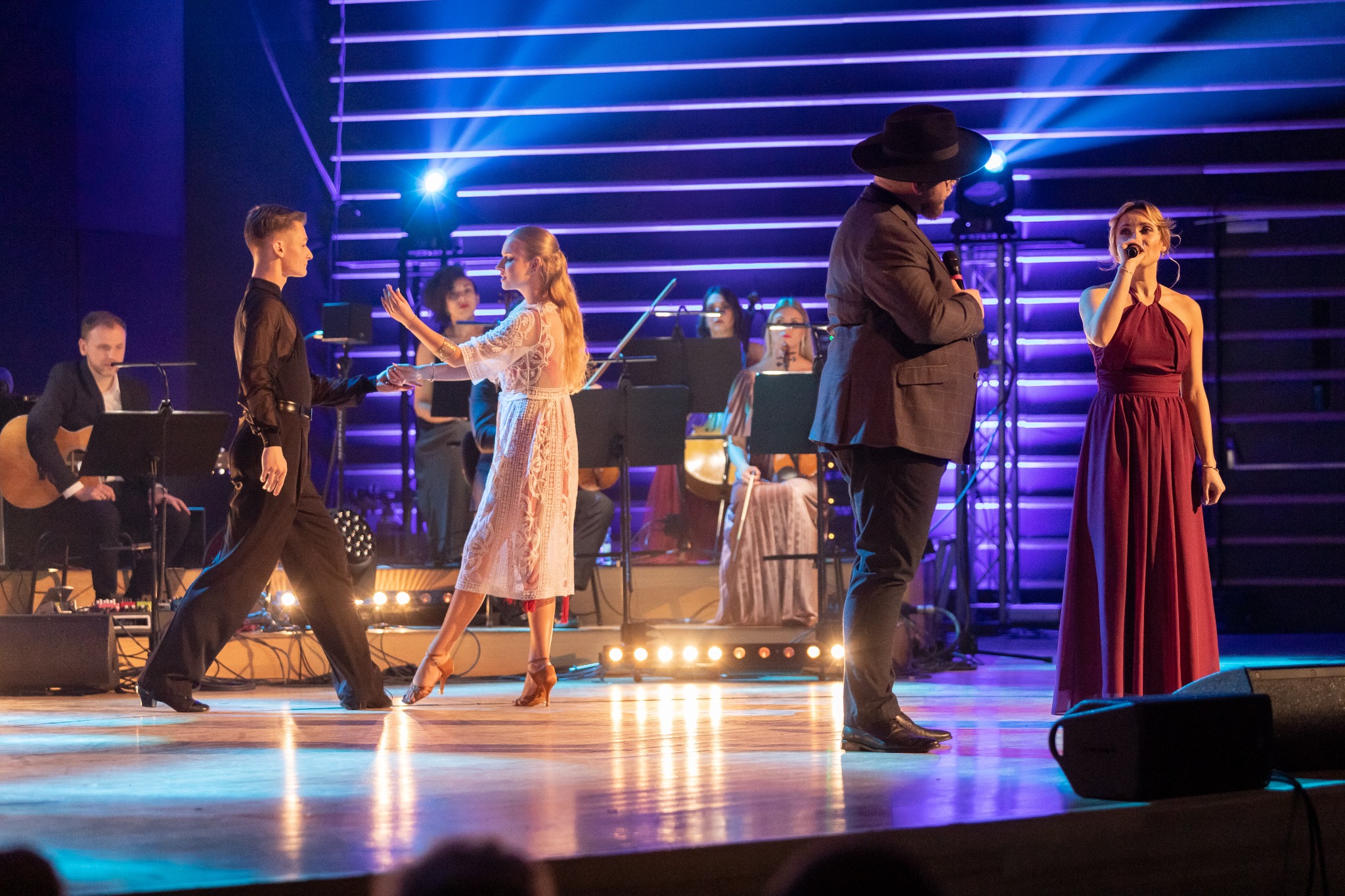Scena z występem na żywo: para taneczna w ruchu, orkiestra w tle, wokalistka w długiej sukni i mężczyzna w kapeluszu z mikrofonem, oświetlenie sceniczne.