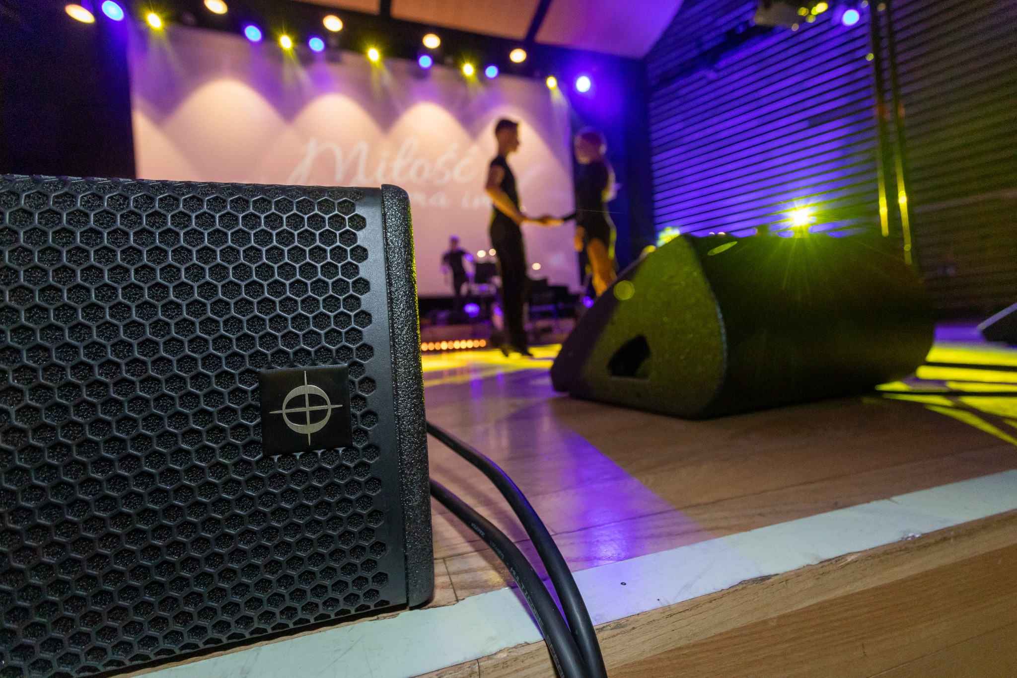 Dwa głośniki na drewnianej scenie, w tle para tancerzy w czarnych strojach pod światłami reflektorów podczas występu.