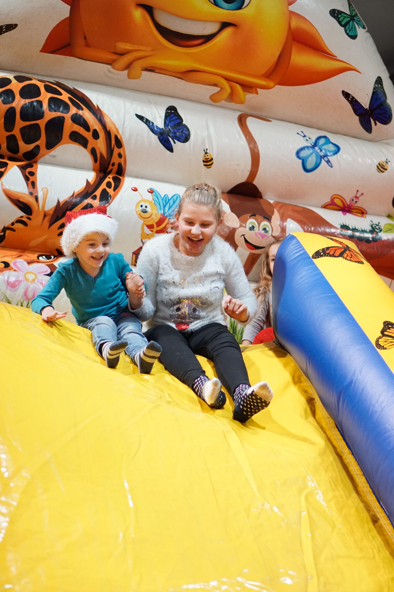 Dwoje roześmianych dzieci zjeżdża z dmuchanej zjeżdżalni z motywami zwierząt i owadów, chłopiec w czapce Mikołaja.