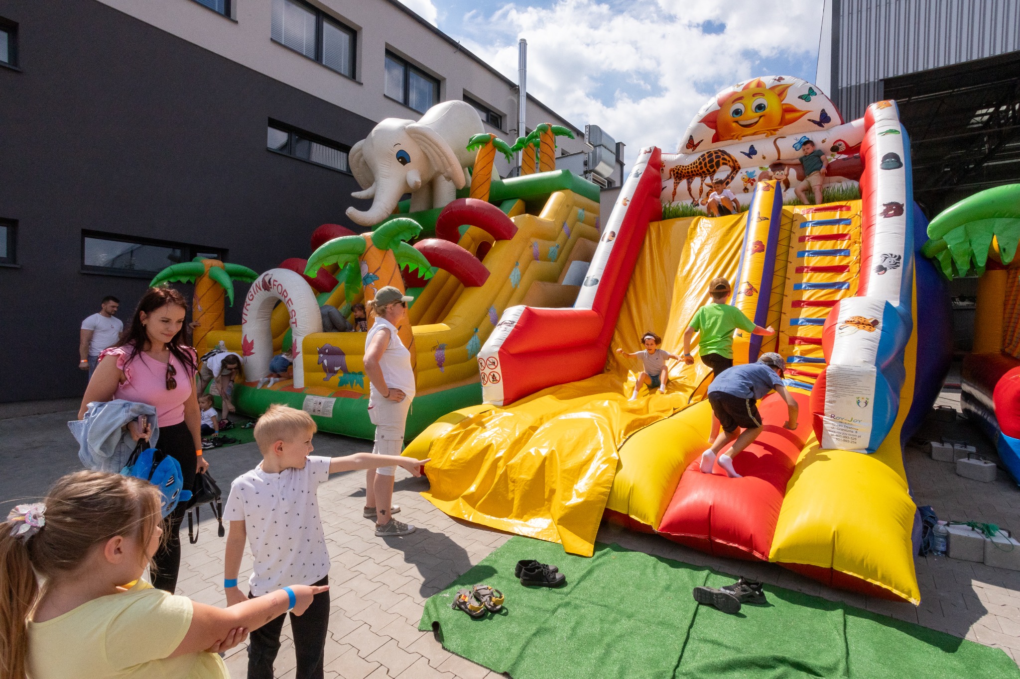 Dzieci bawią się na dmuchanym zamku z motywami zwierzęcymi i palmami, ustawionym na zewnątrz budynku w słoneczny dzień.