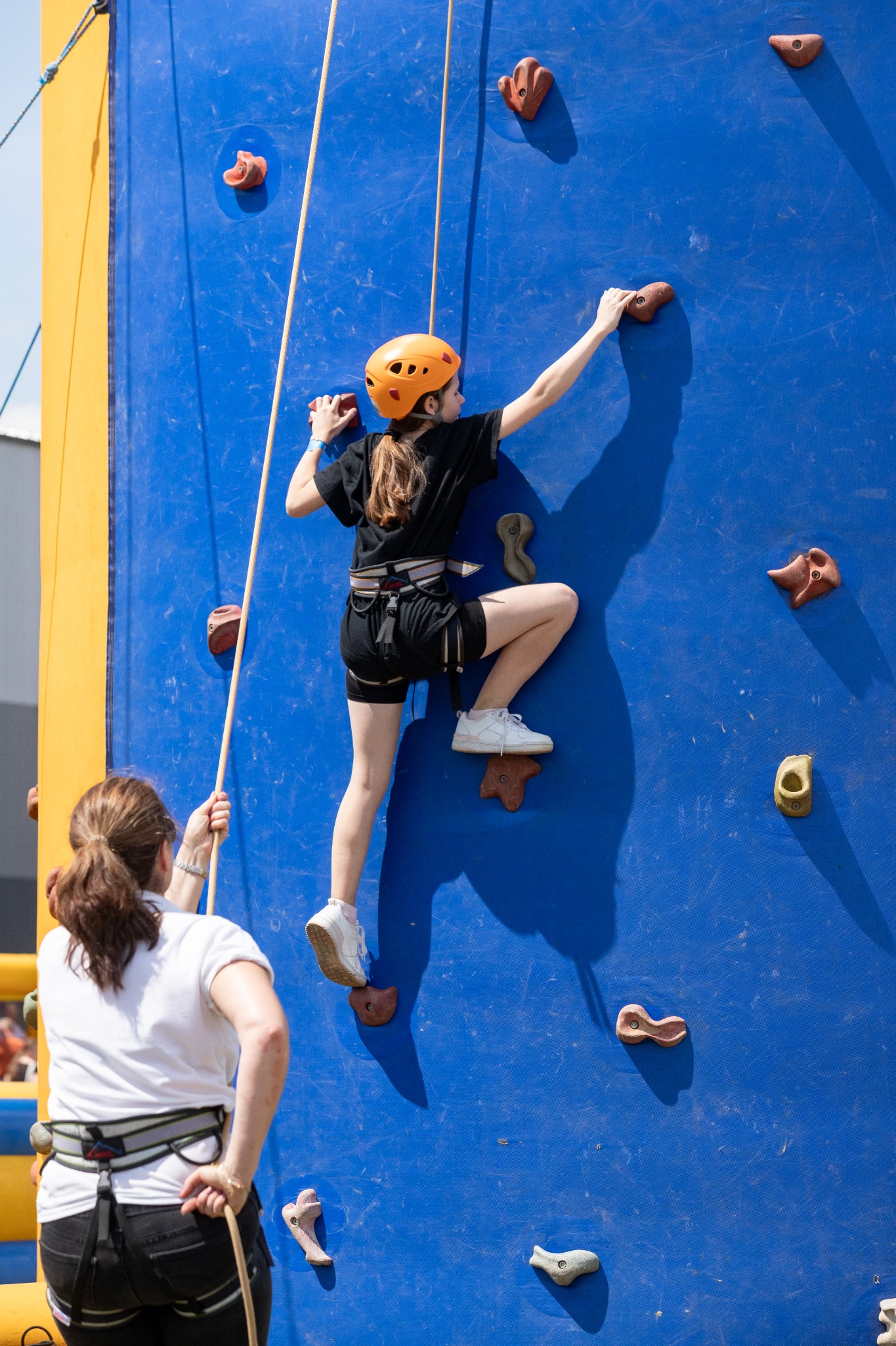 Dziewczynka w pomarańczowym kasku wspina się na niebieską ściankę wspinaczkową z różnokolorowymi uchwytami, zabezpieczana liną przez instruktorkę w białej koszulce.
