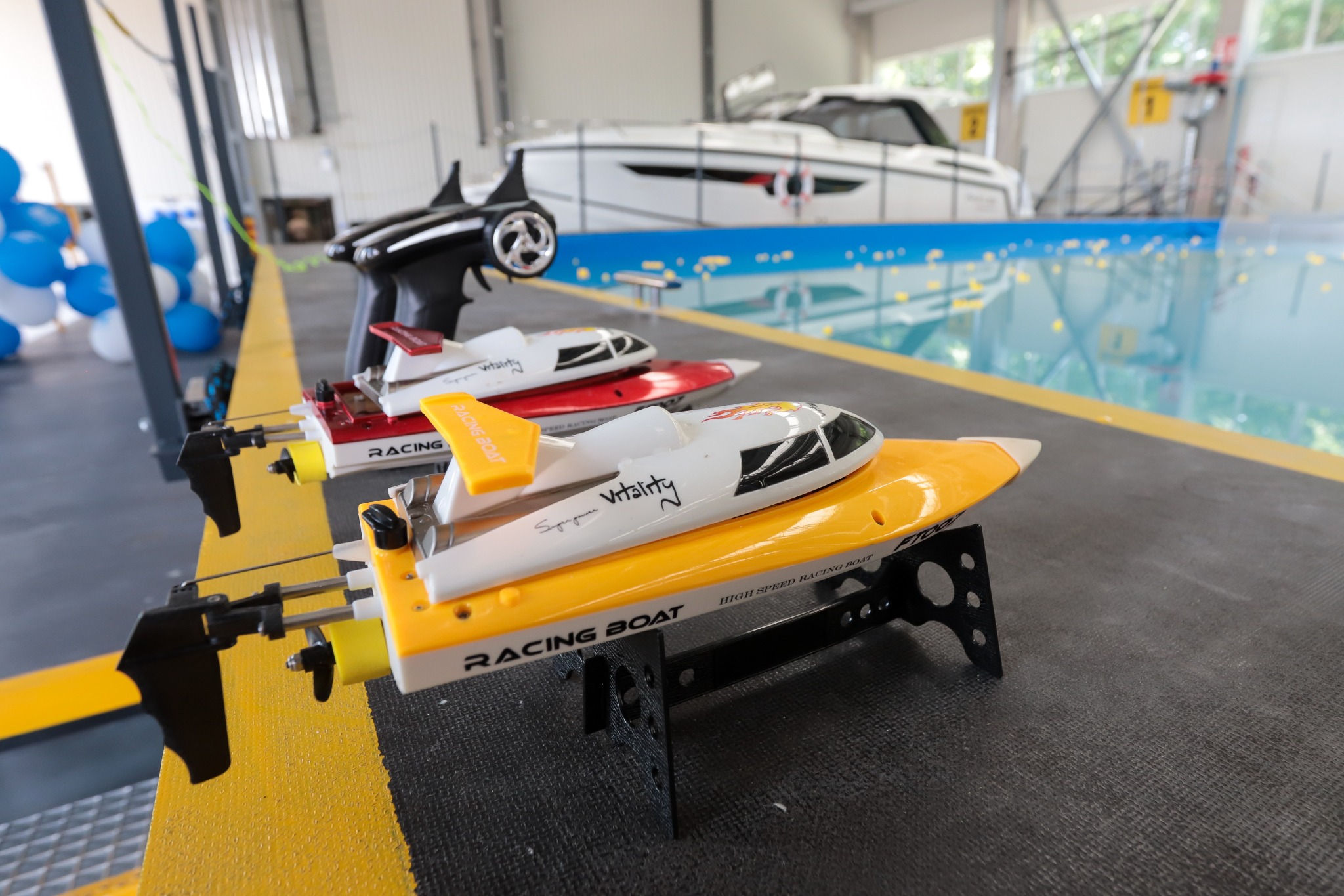 Trzy modele zdalnie sterowanych łodzi wyścigowych na tle basenu i dużej łodzi motorowej. Żółta łódź na pierwszym planie z napisem 'Racing Boat'.