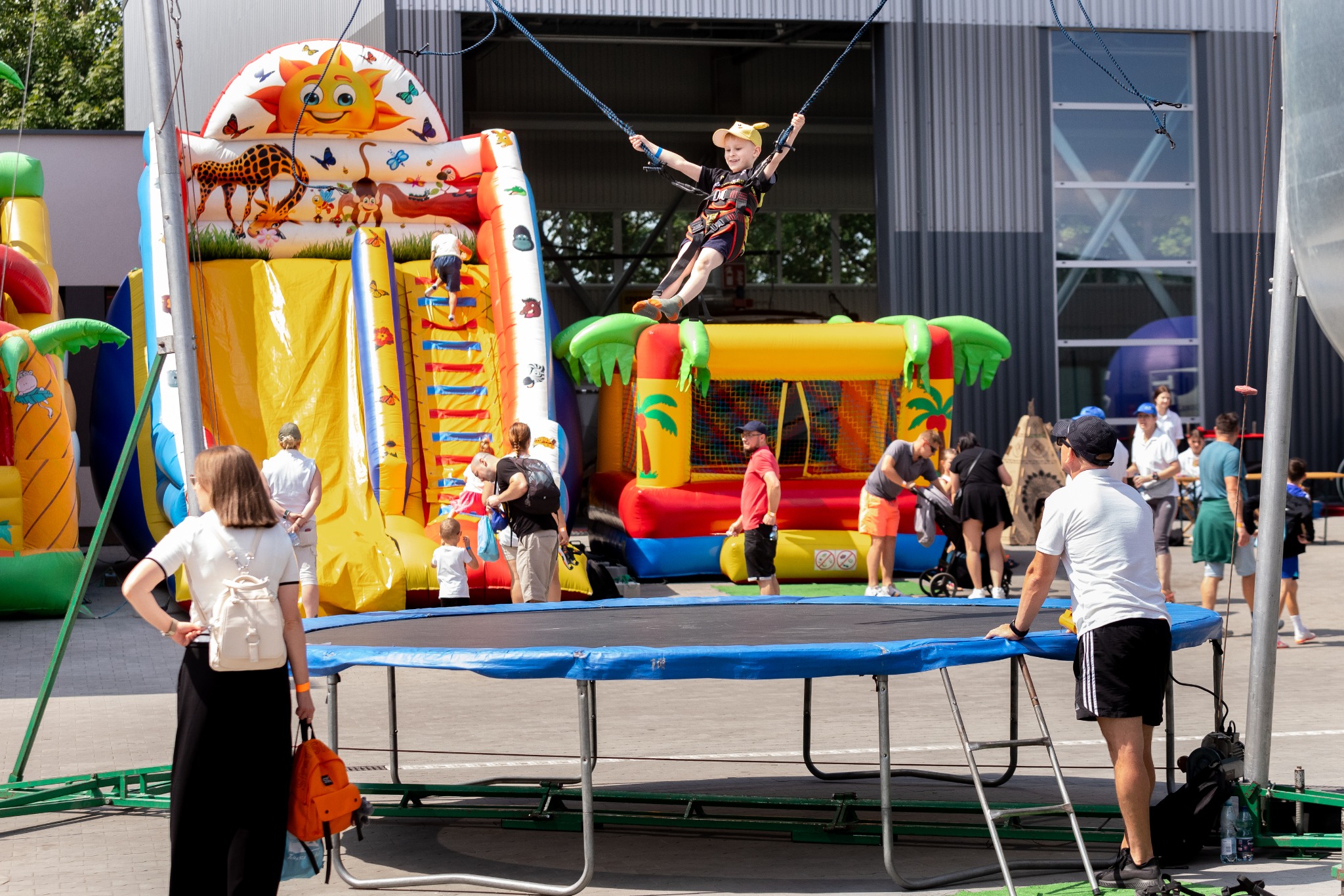 Chłopiec w uprzęży skacze na trampolinie bungee na tle dmuchanych zamków i zjeżdżalni podczas imprezy plenerowej.