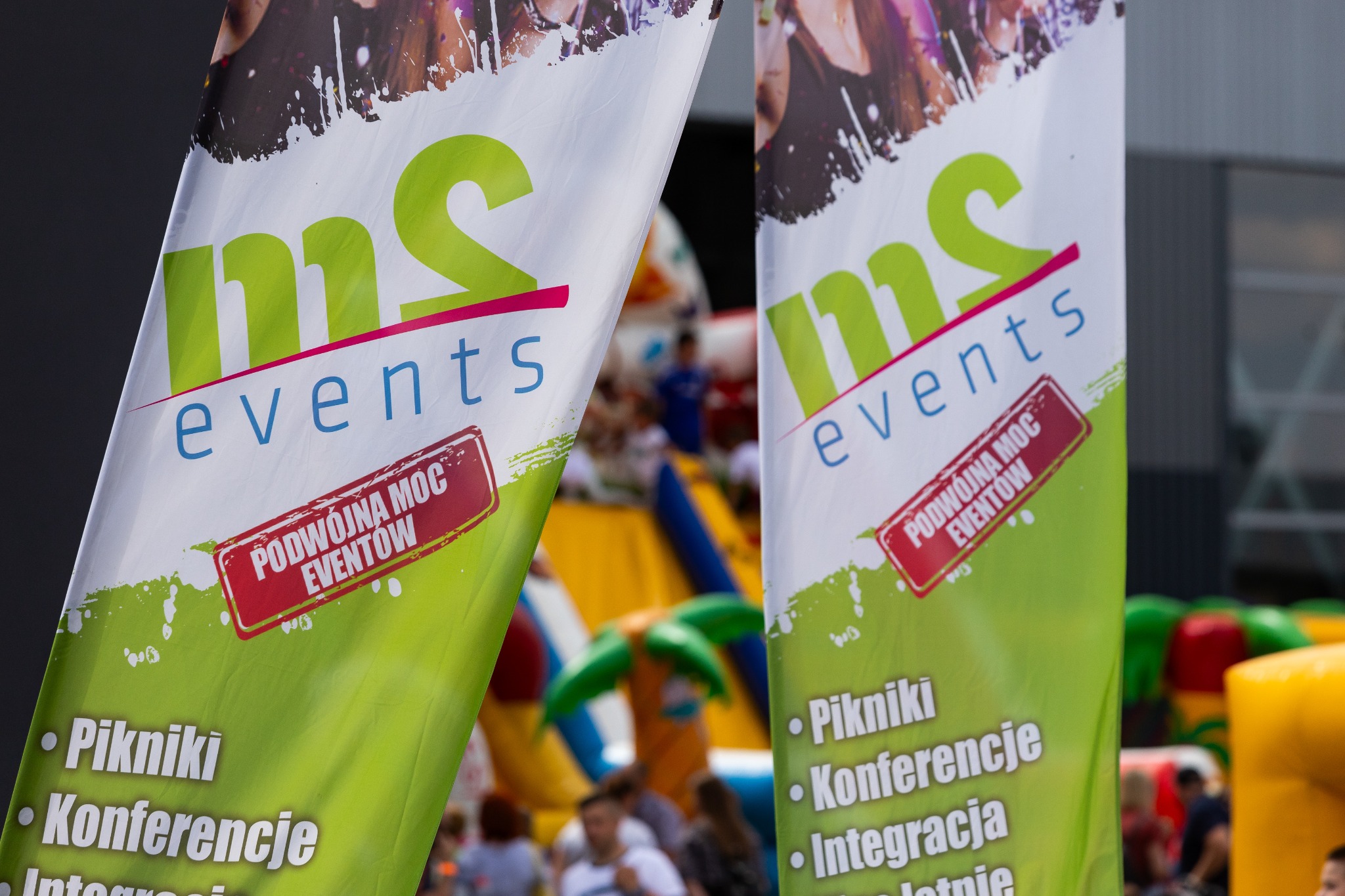 Dwa banery reklamowe z logo 'ms events' i hasłem 'Podwójna Moc Eventów', na tle rozmytego dmuchanego placu zabaw i grupy ludzi.