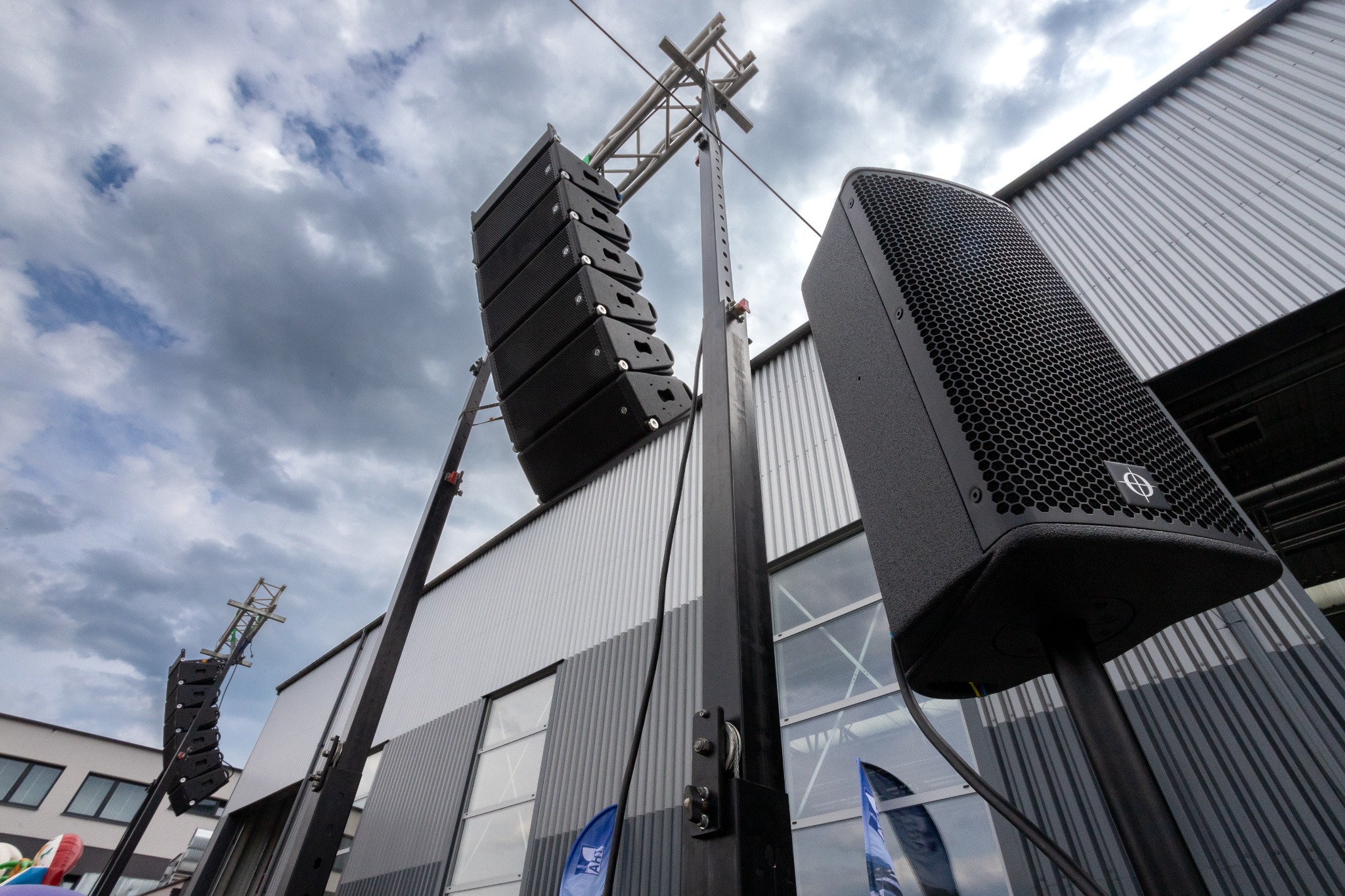 Zestaw nagłośnieniowy line array i pojedynczy głośnik na statywach, ustawione na zewnątrz budynku o szarej, metalowej fasadzie, pod pochmurnym niebem.