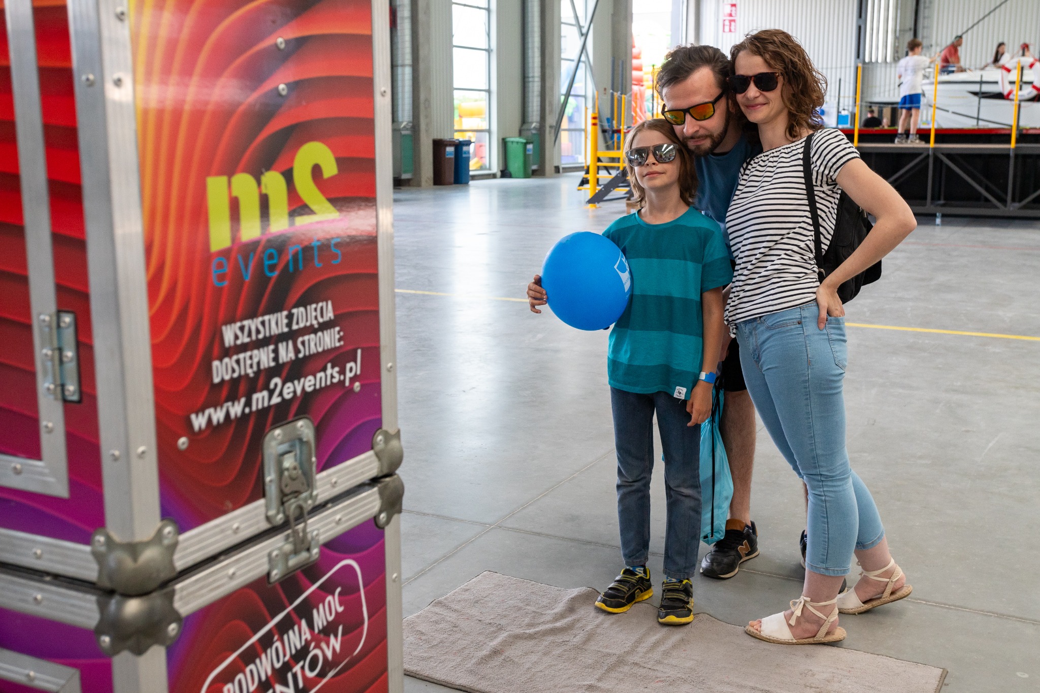 Rodzina pozuje obok skrzyni transportowej z logo firmy eventowej, w tle widoczna konstrukcja sceniczna i dmuchany zamek.