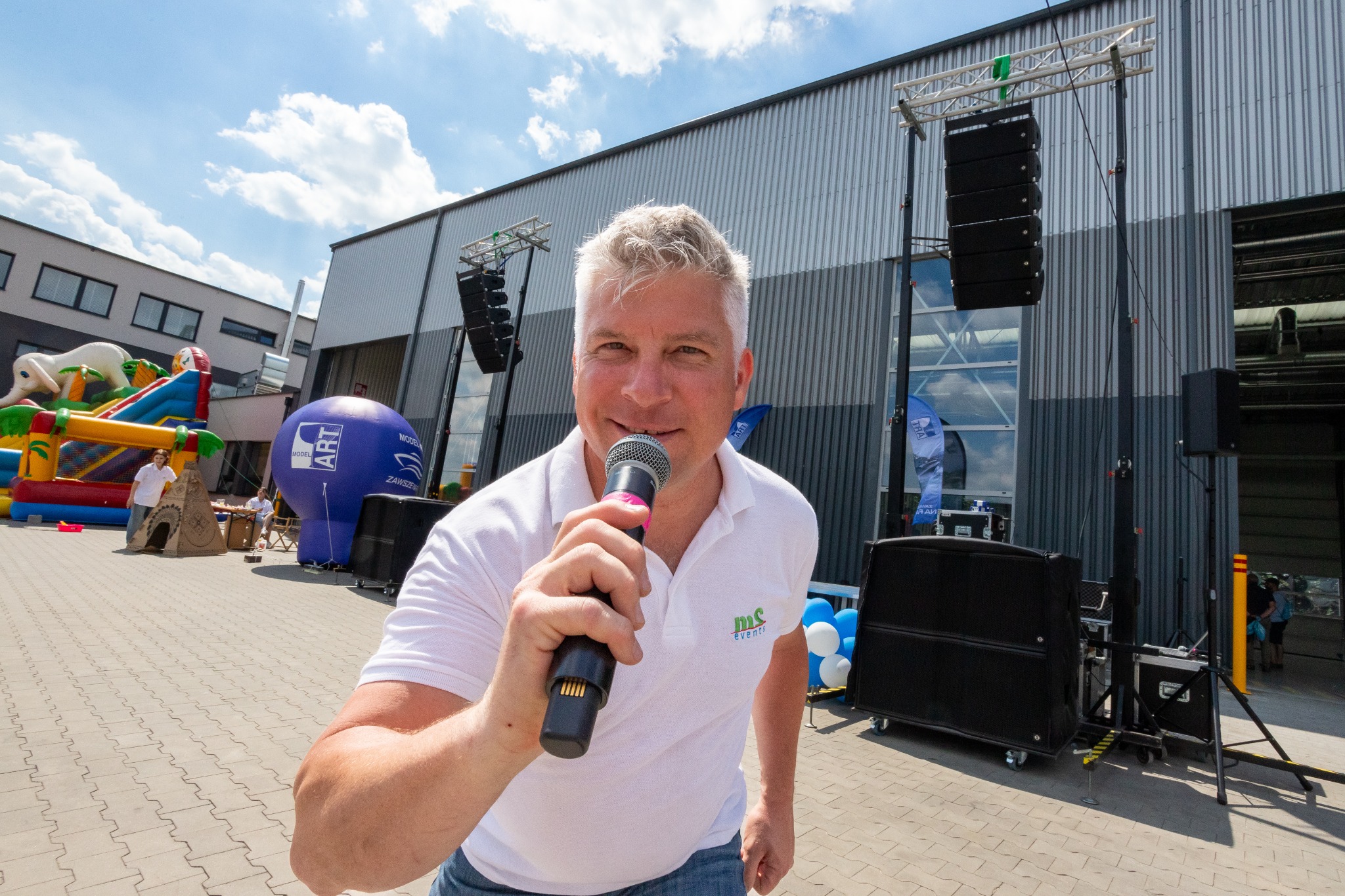 Uśmiechnięty mężczyzna z mikrofonem na tle sceny z nagłośnieniem, dmuchańcami i balonami reklamowymi.