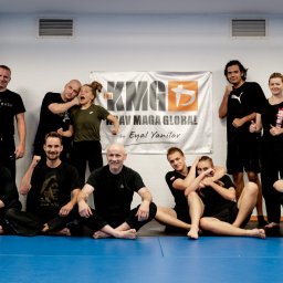 Krav Maga Silesia - Grupa osób po treningu Krav Maga Global, siedząca i stojąca na niebieskiej macie, za nimi flaga z logo KMG i nazwiskiem Eyal Yanilov.