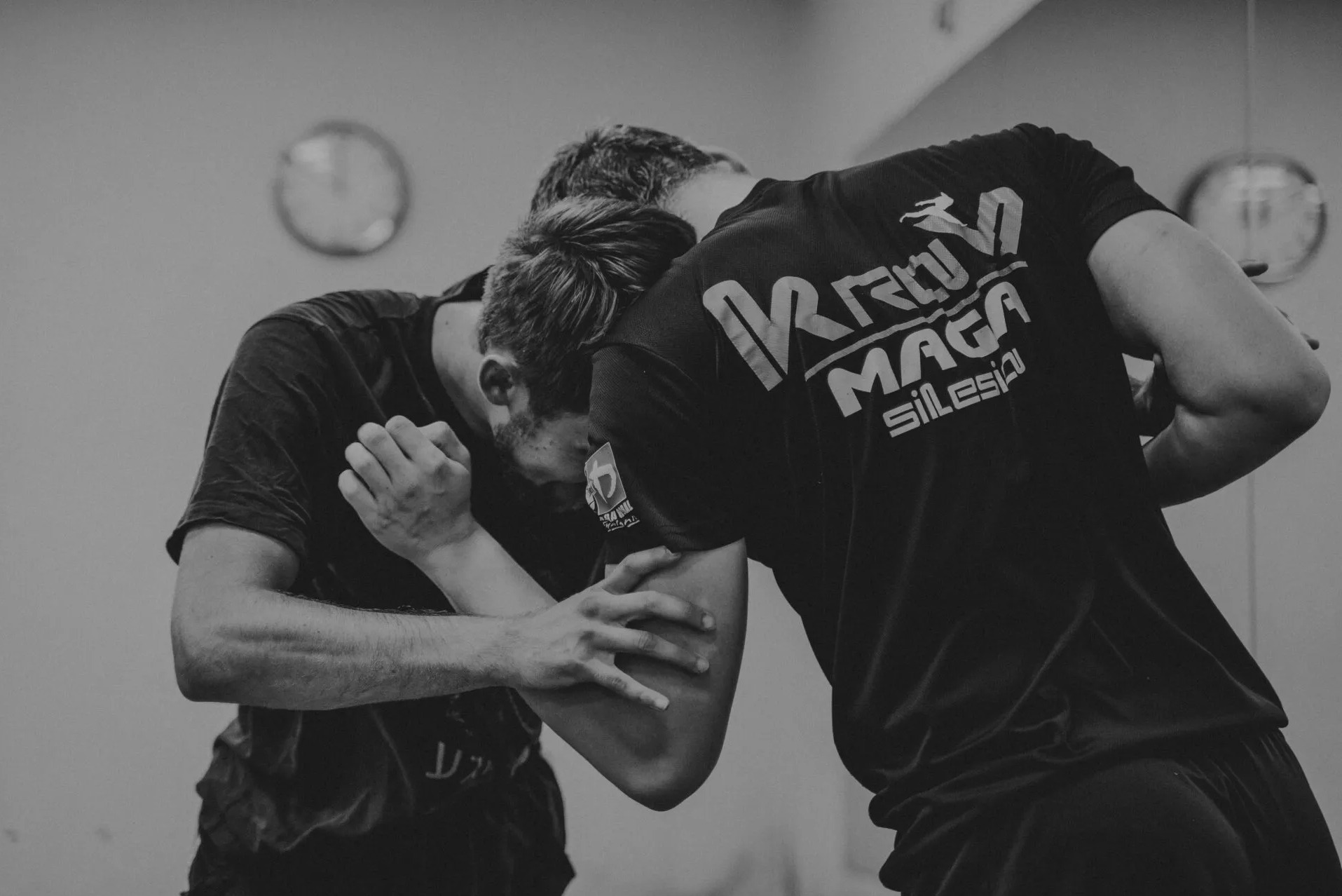 Czarno-białe zdjęcie dwóch mężczyzn w trakcie sparingu, jeden z nich ma na koszulce napis 'Krav Maga Silesia', w tle widoczne zegary na ścianie.