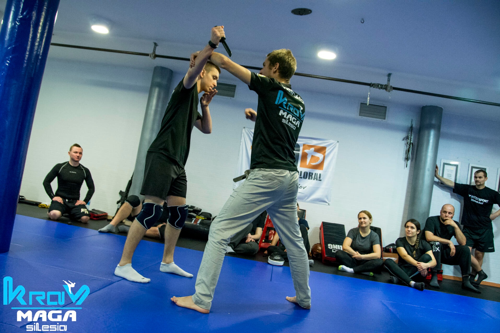 Trening Krav Maga: instruktor demonstruje obronę przed atakiem nożem, uczestnik w blokującej pozycji, na niebieskiej macie, w tle grupa obserwujących i logo Krav Maga Silesia.