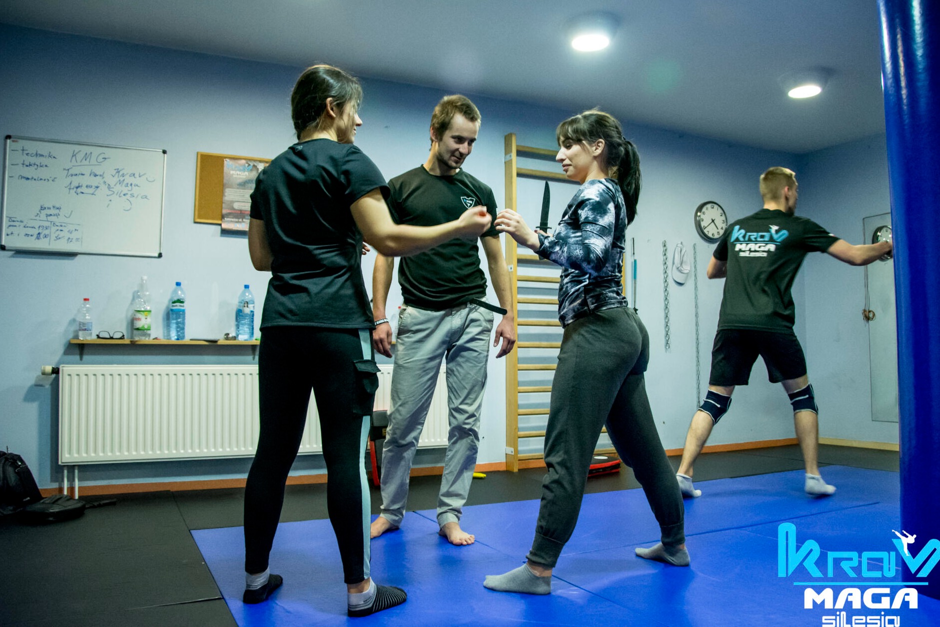 Instruktor demonstruje technikę obrony przed nożem na sali treningowej Krav Maga, z uczestnikami w tle i tablicą z notatkami na ścianie.
