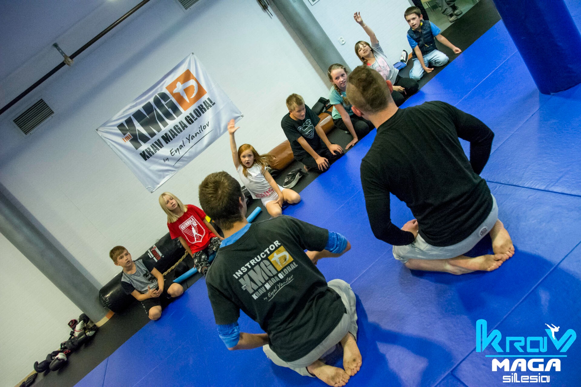 Instruktor Krav Maga prowadzi zajęcia dla grupy dzieci i młodzieży na niebieskiej macie, w tle widoczny baner KMG Krav Maga Global.