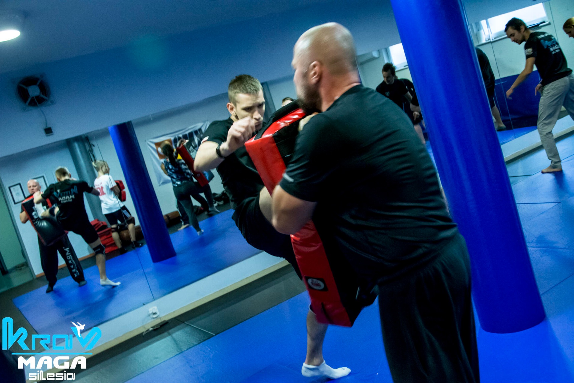 Mężczyzna trenuje uderzenie kolanem w tarczę trzymaną przez trenera podczas zajęć grupowych w sali z lustrami i niebieską podłogą.