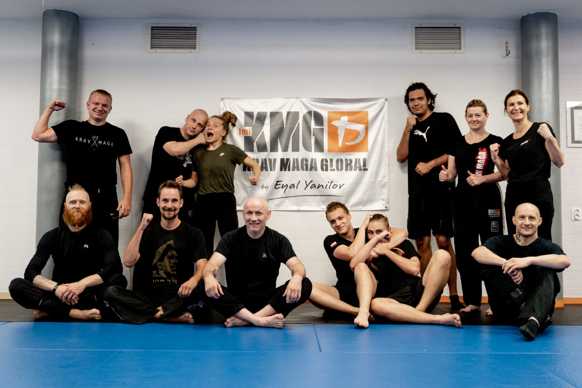 Grupa osób po treningu Krav Maga Global, siedząca i stojąca na niebieskiej macie, za nimi flaga z logo KMG i nazwiskiem Eyal Yanilov.