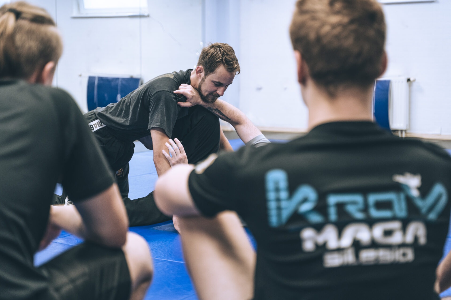 Mężczyzna z brodą w czarnej koszulce wykonuje dźwignię na ramieniu drugiego mężczyzny, na niebieskiej macie, podczas zajęć grupowych.