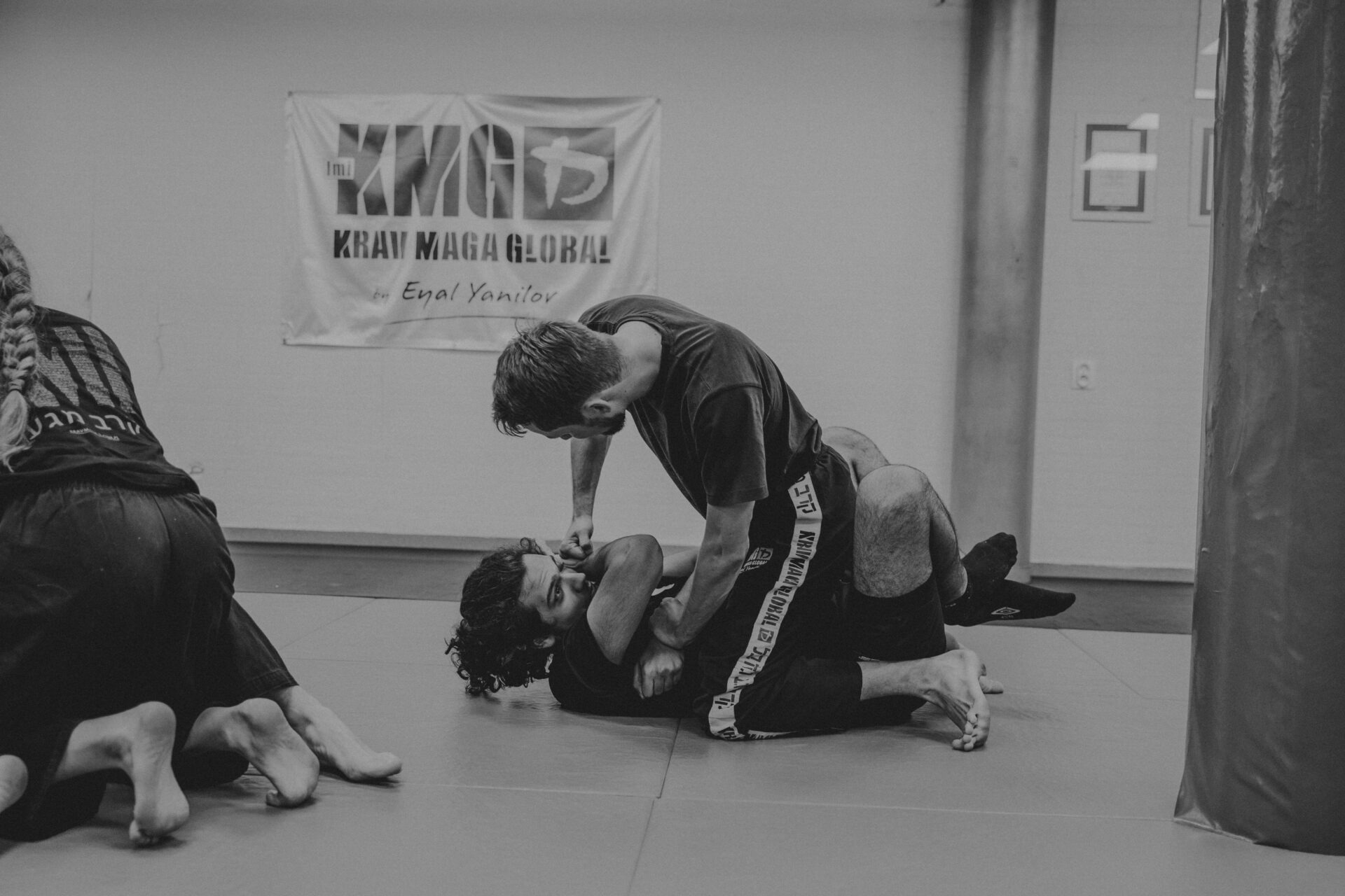 Czarno-białe zdjęcie przedstawia dwóch mężczyzn ćwiczących Krav Maga na macie w sali treningowej, z logo Krav Maga Global widocznym w tle.