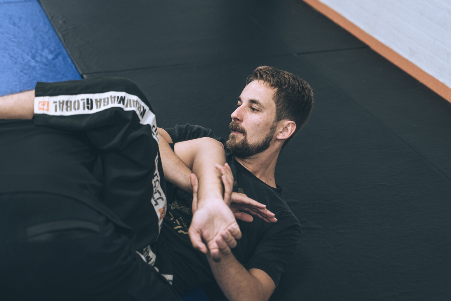 Mężczyzna z brodą w czarnej koszulce demonstruje technikę samoobrony, leżąc na macie z innym mężczyzną w spodniach z napisem 'KRAV MAGA GLOBAL'.