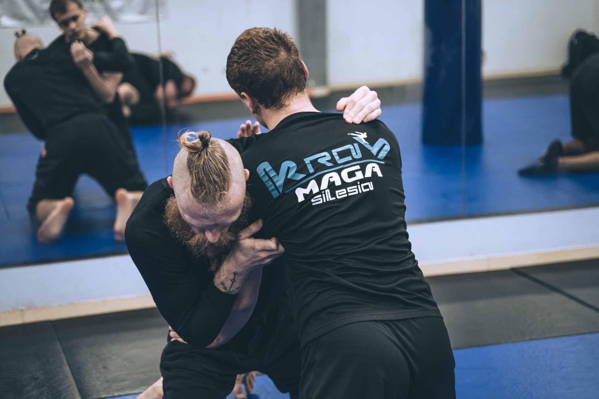 Dwóch mężczyzn w czarnych strojach ćwiczy chwyt na macie, widoczne odbicie w lustrze i logo 'Krav Maga Silesia' na koszulce jednego z nich.