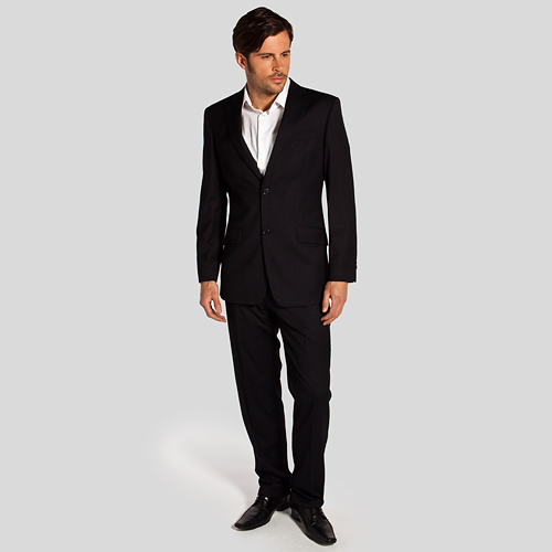 Mężczyzna w czarnym, eleganckim garniturze i białej koszuli, pozujący na jasnoszarym tle, z delikatnym spojrzeniem w bok.